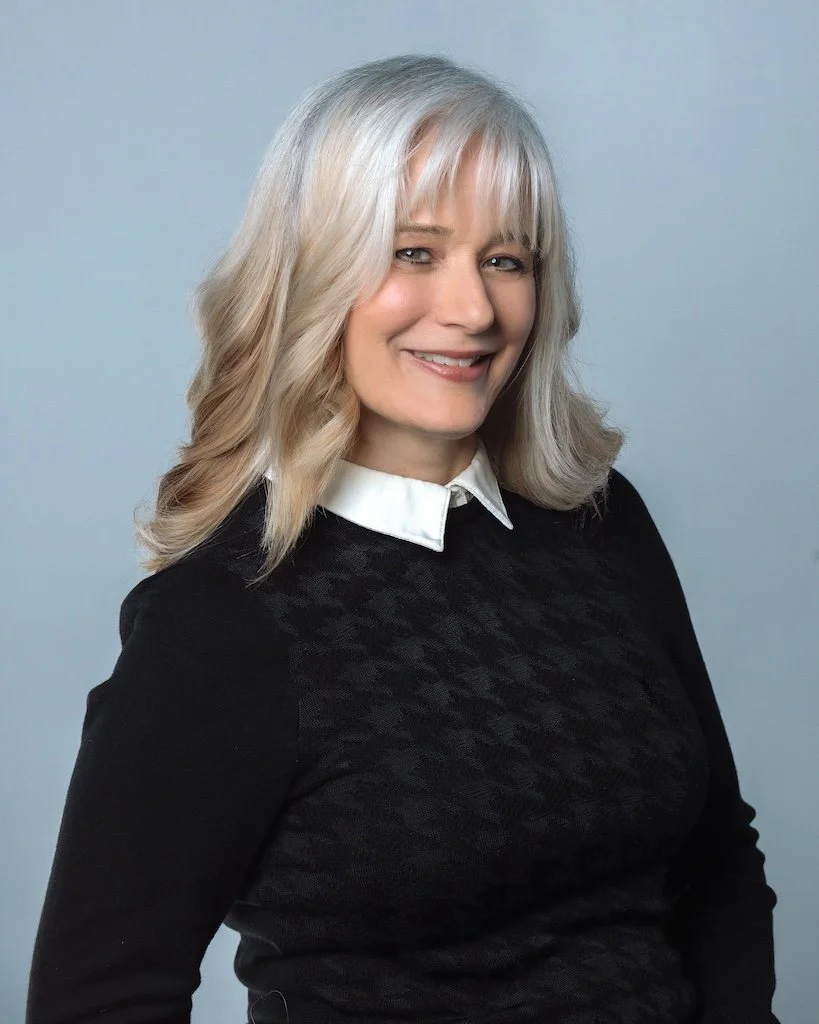 A woman with shoulder-length blonde hair smiling at the camera, wearing a black sweater with a white collared shirt underneath, against a light blue background.