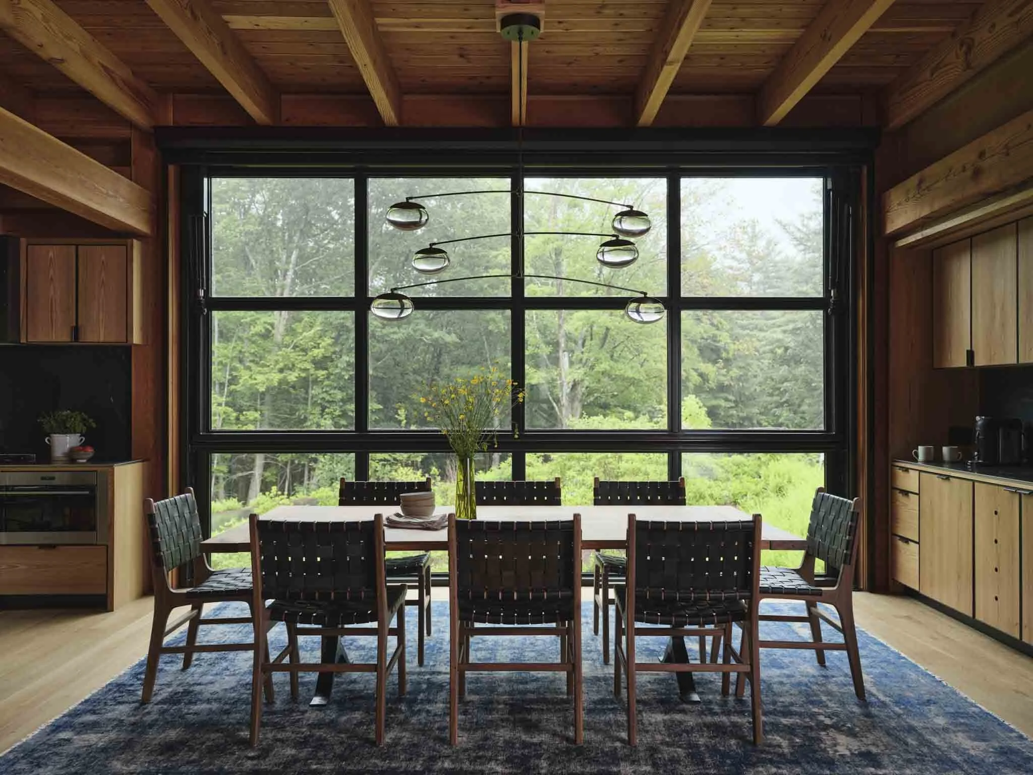 A dining room with large windows revealing a green forest outside, a wooden dining table with eight chairs, a blue patterned rug, and a modern chandelier hanging above the table.