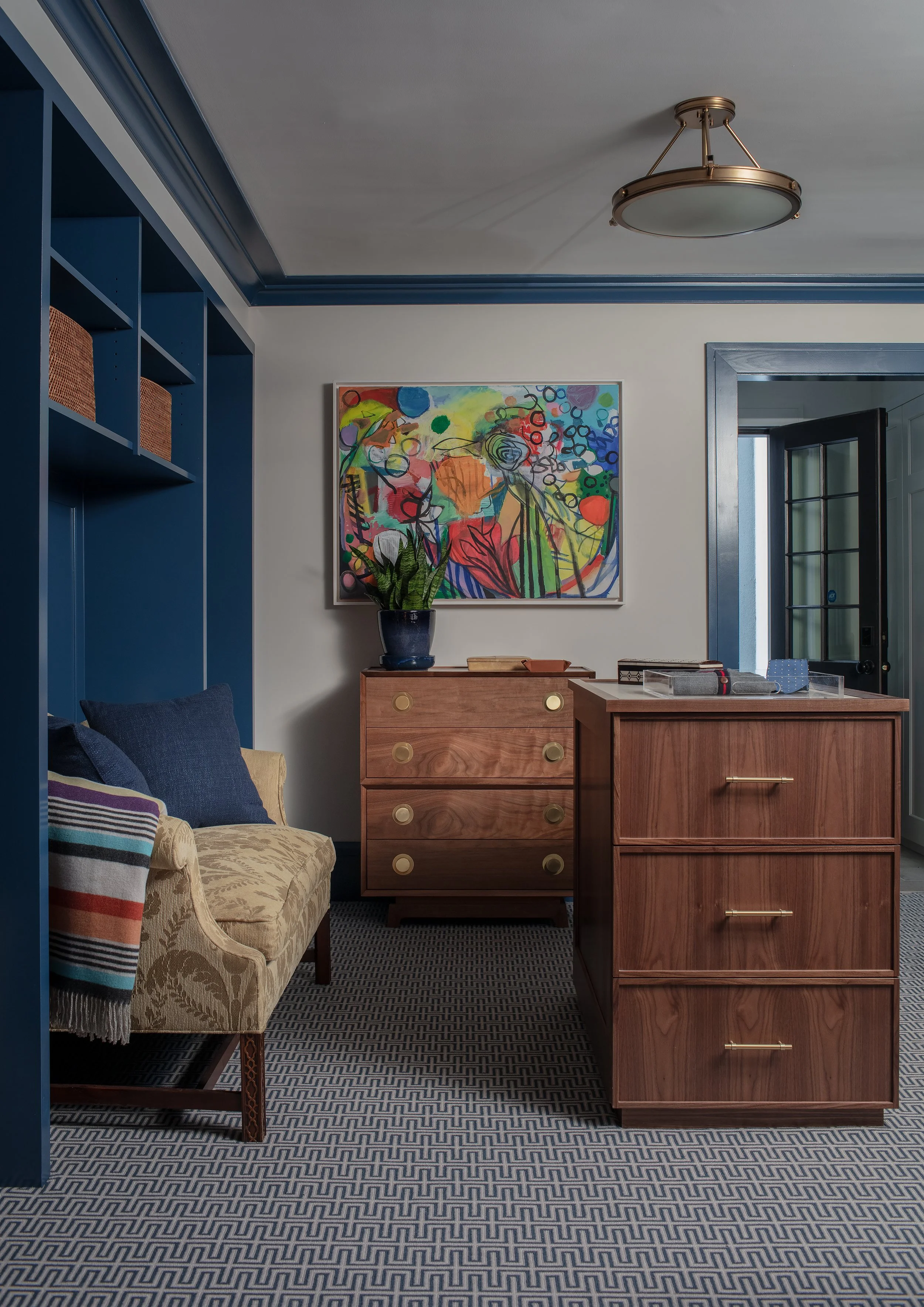 Interior of a room with a beige patterned sofa with colorful striped blanket, blue pillows, blue shelves, a wooden chest of drawers with a potted plant, colorful abstract painting, and a wooden cabinet. Dark blue trim and door frame, ceiling light fixture, patterned carpet.
