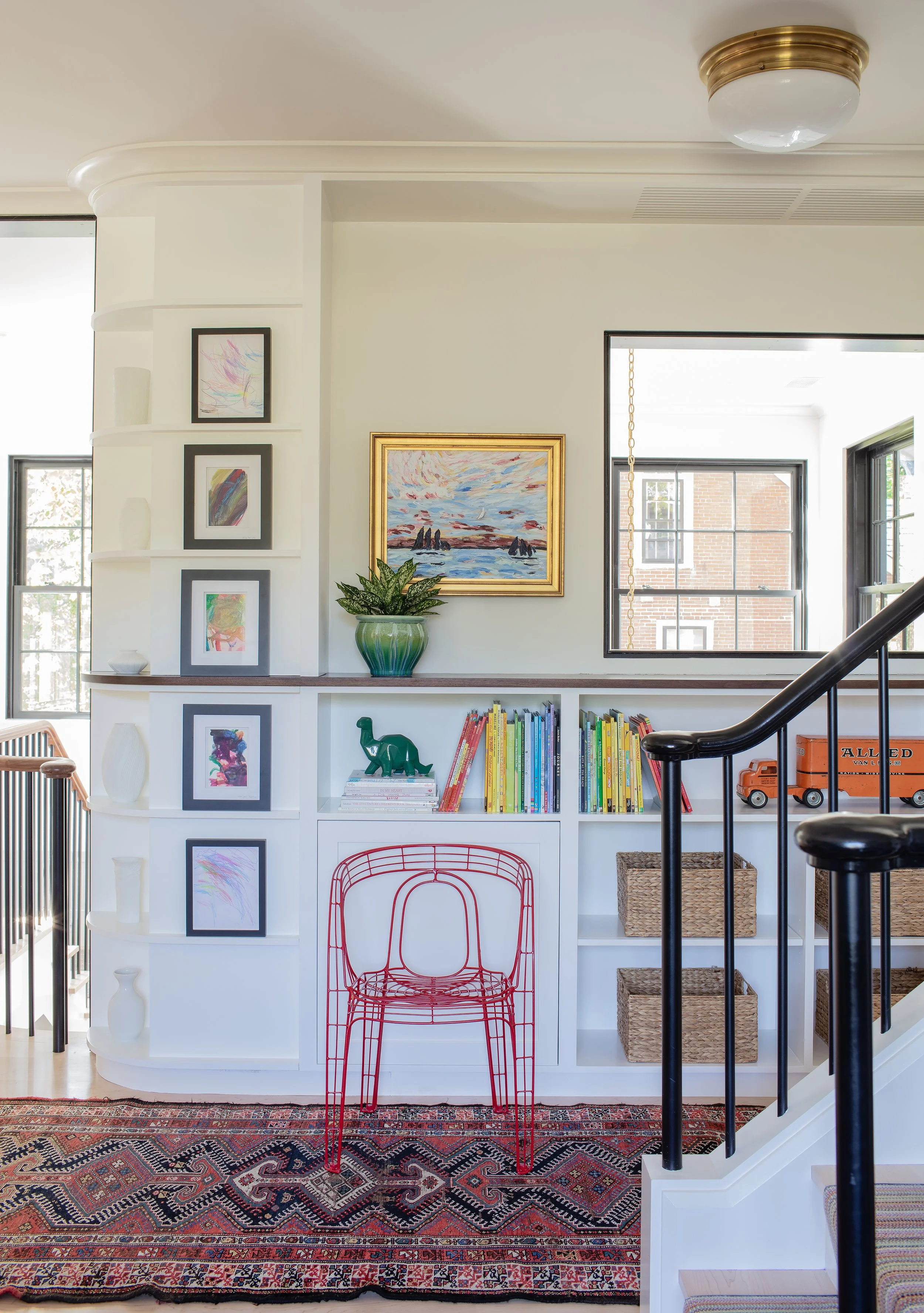 Interior view of a living space with white built-in shelves, framed artwork, and a red wire chair, with a patterned rug and black stair railing.