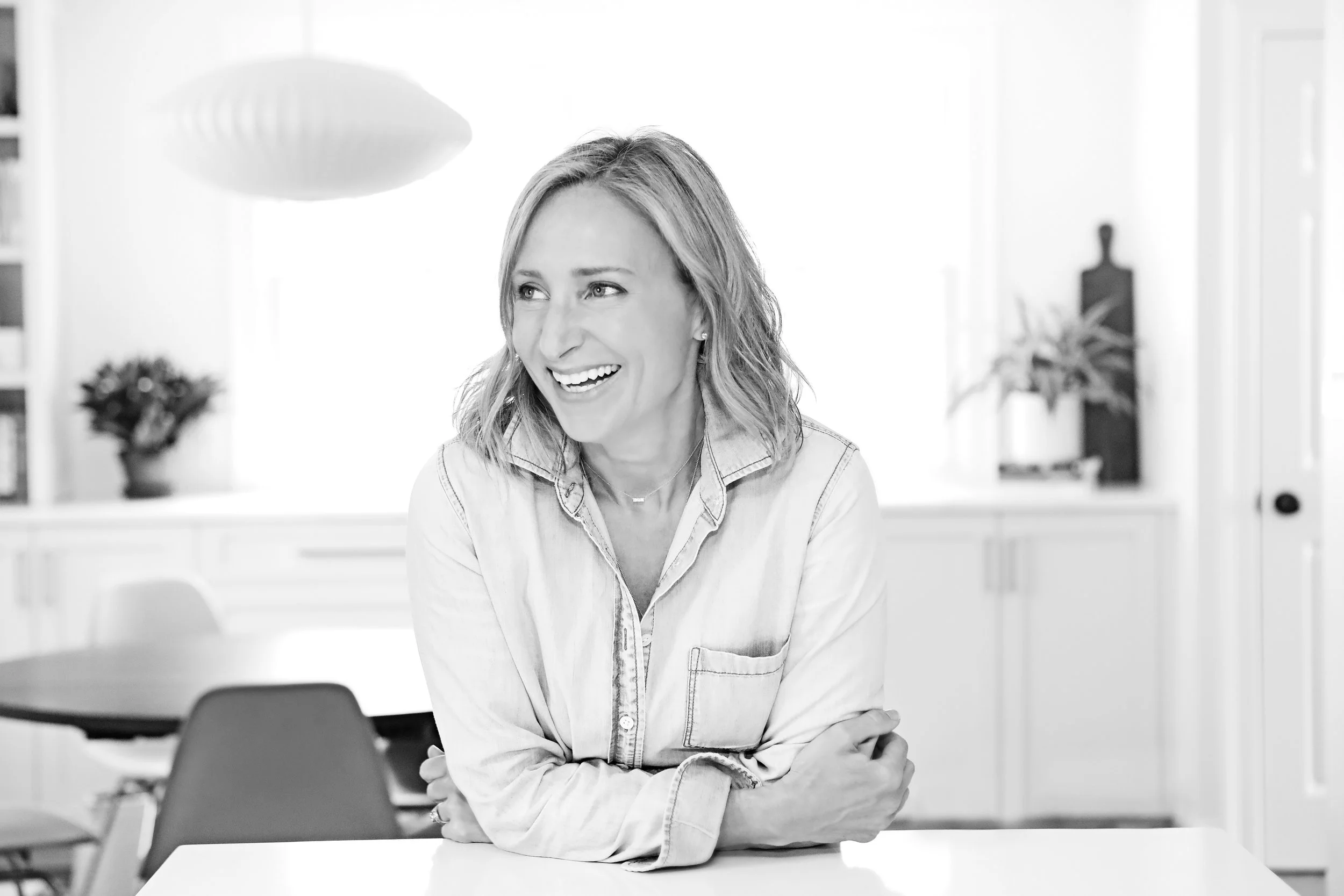 A woman with shoulder-length hair smiling and sitting at a table in a bright, modern kitchen.