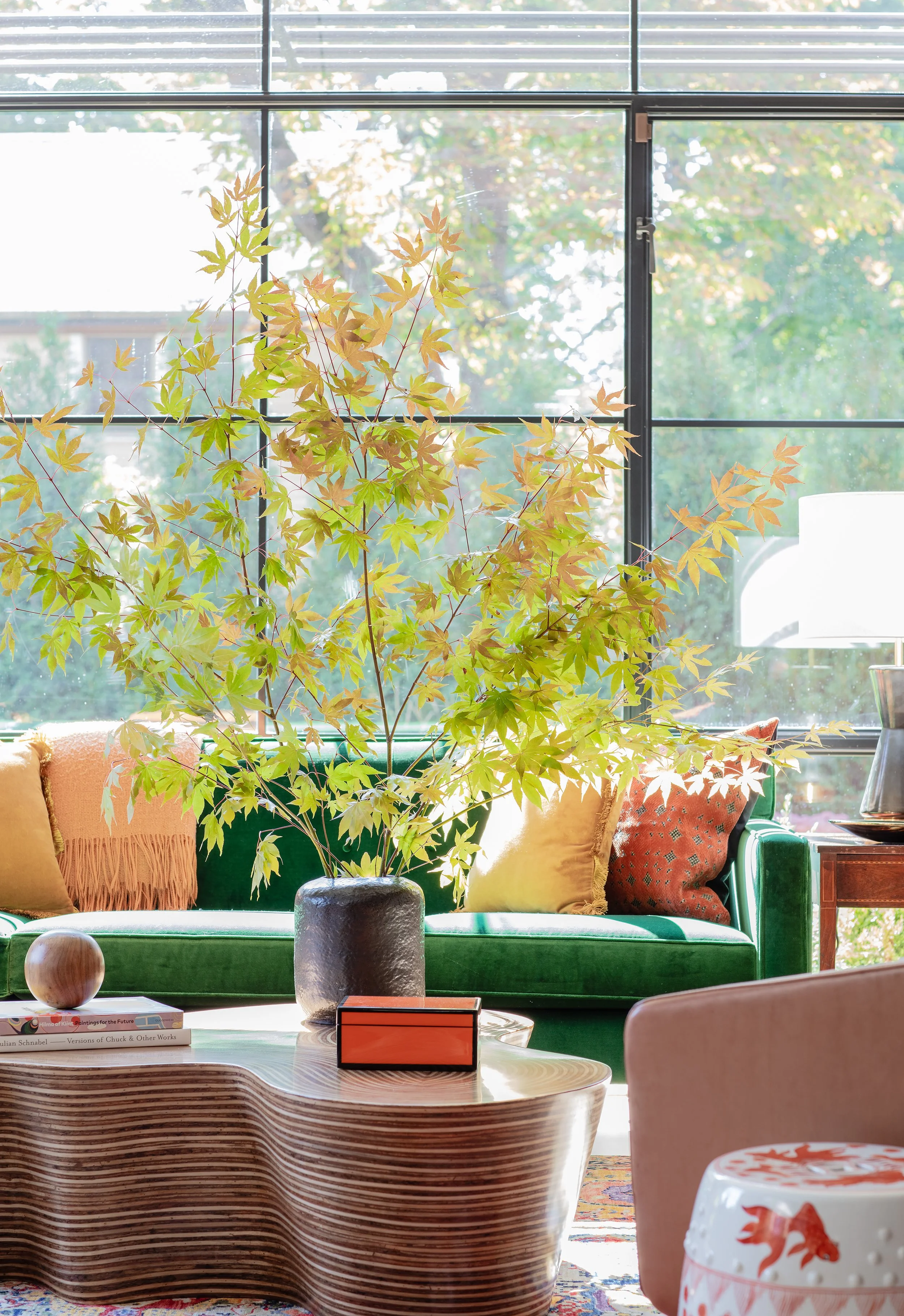 Living room with a green velvet sofa, colorful pillows, a wooden coffee table with books and decorative items, a large plant in a black vase, and a window with trees outside.