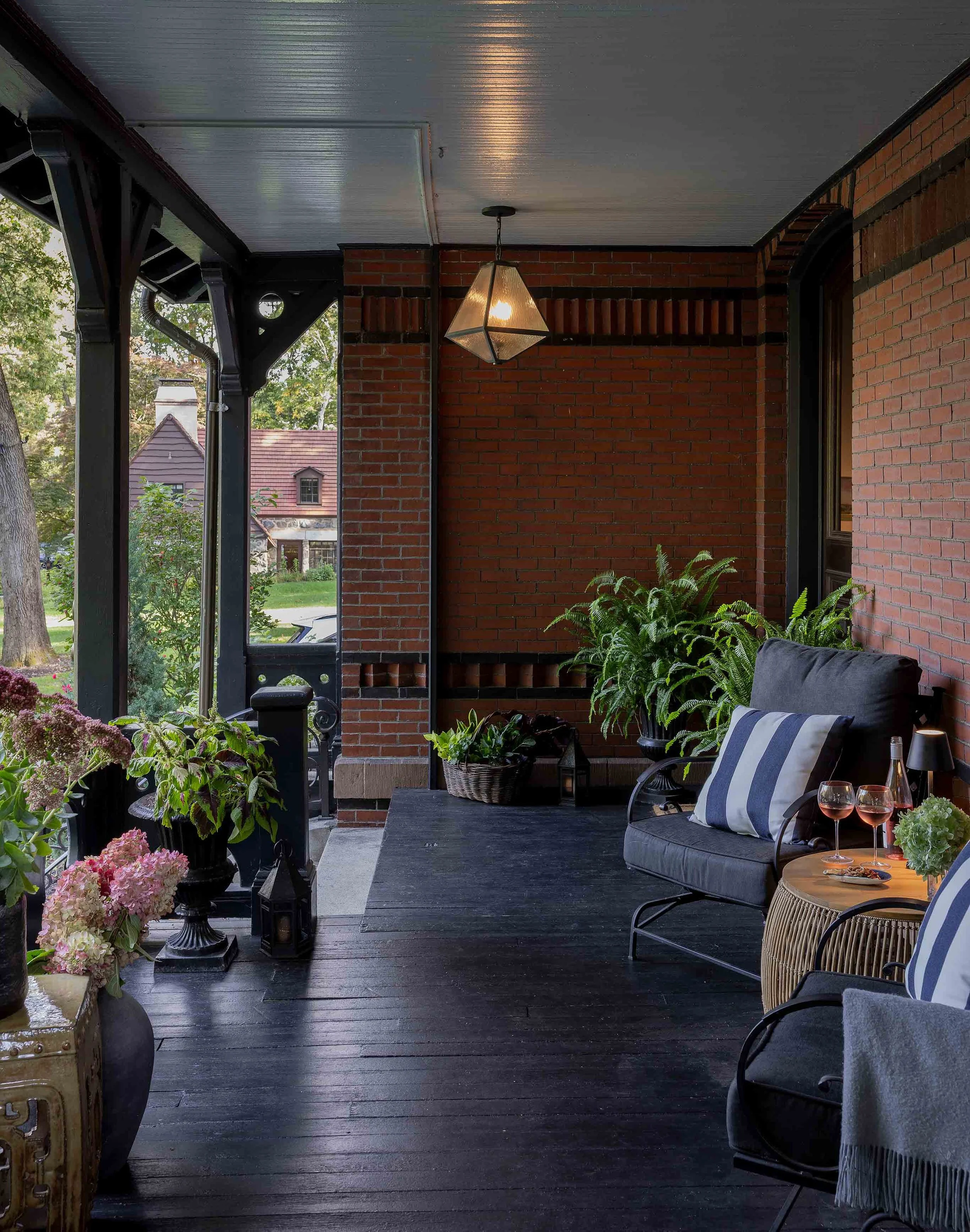 A cozy, covered porch with brick walls and black wooden flooring. It features black metal chairs with cushions, a small round table with two glasses of pink wine, and lush green plants and potted flowers along the railing and in baskets. A pendant light hangs from the ceiling, and residential houses are visible in the background.