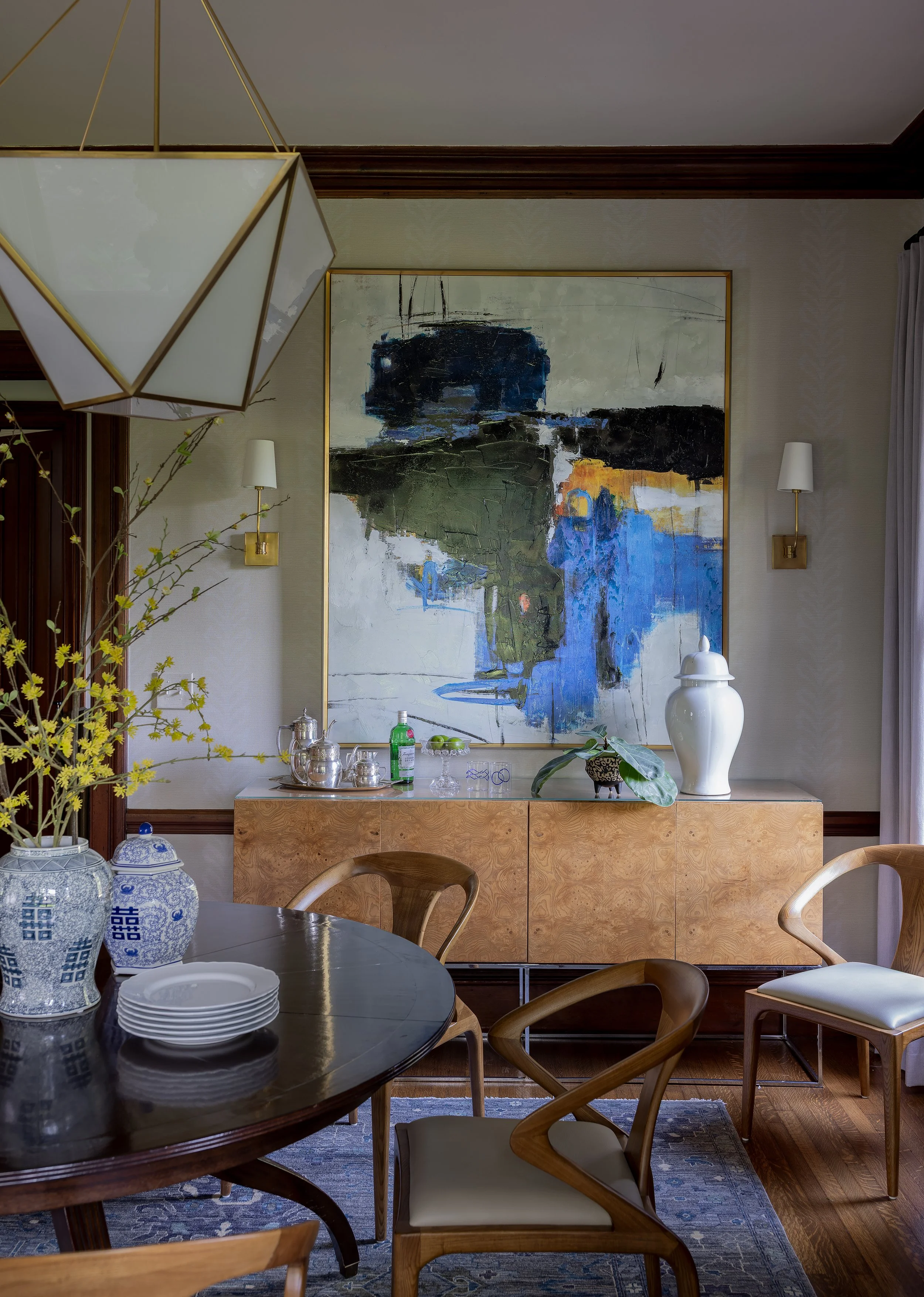 A dining room with a wooden round table, chairs, and decorative items including vases, a large abstract painting, and a sideboard with glassware and a white ceramic vase.