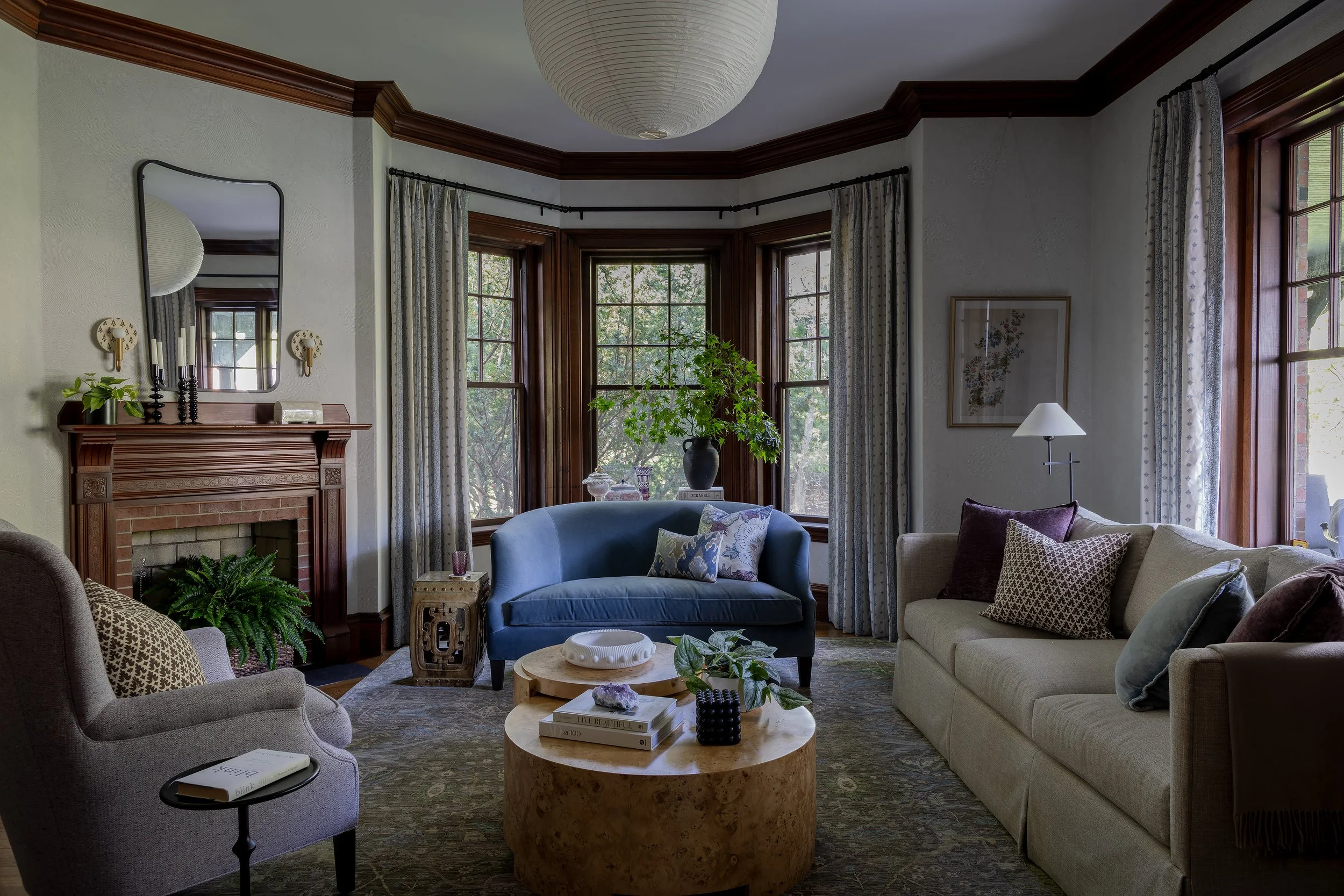 Living room with large windows, a fireplace with a wooden mantel, a mirror above the mantel, a beige sofa with throw pillows, a blue loveseat, a small side table, a round stone coffee table with books and decorative items, a floor lamp, and curtains with a patterned design.