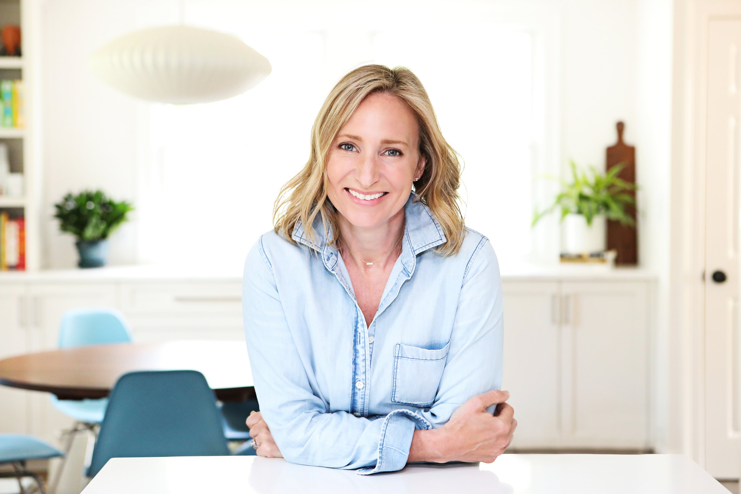 A woman with shoulder-length blonde hair, smiling, wearing a light denim shirt, sitting at a white table in a bright, modern kitchen with white cabinets and green plants.