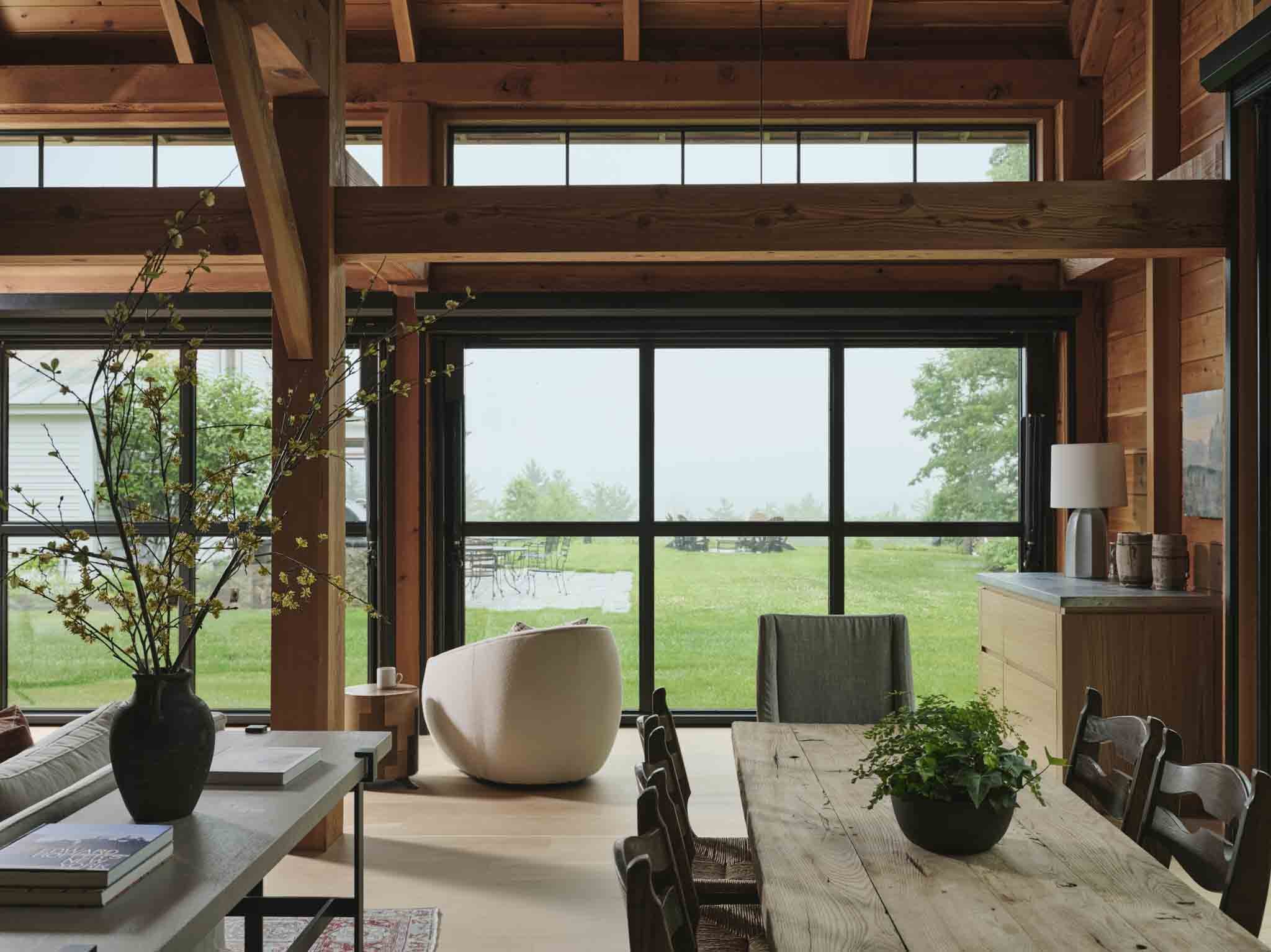Interior of a wooden house living room with large windows looking out to a green yard, a dining table with chairs, a white armchair, a sideboard with decor, and a large vase with branches on a side table.