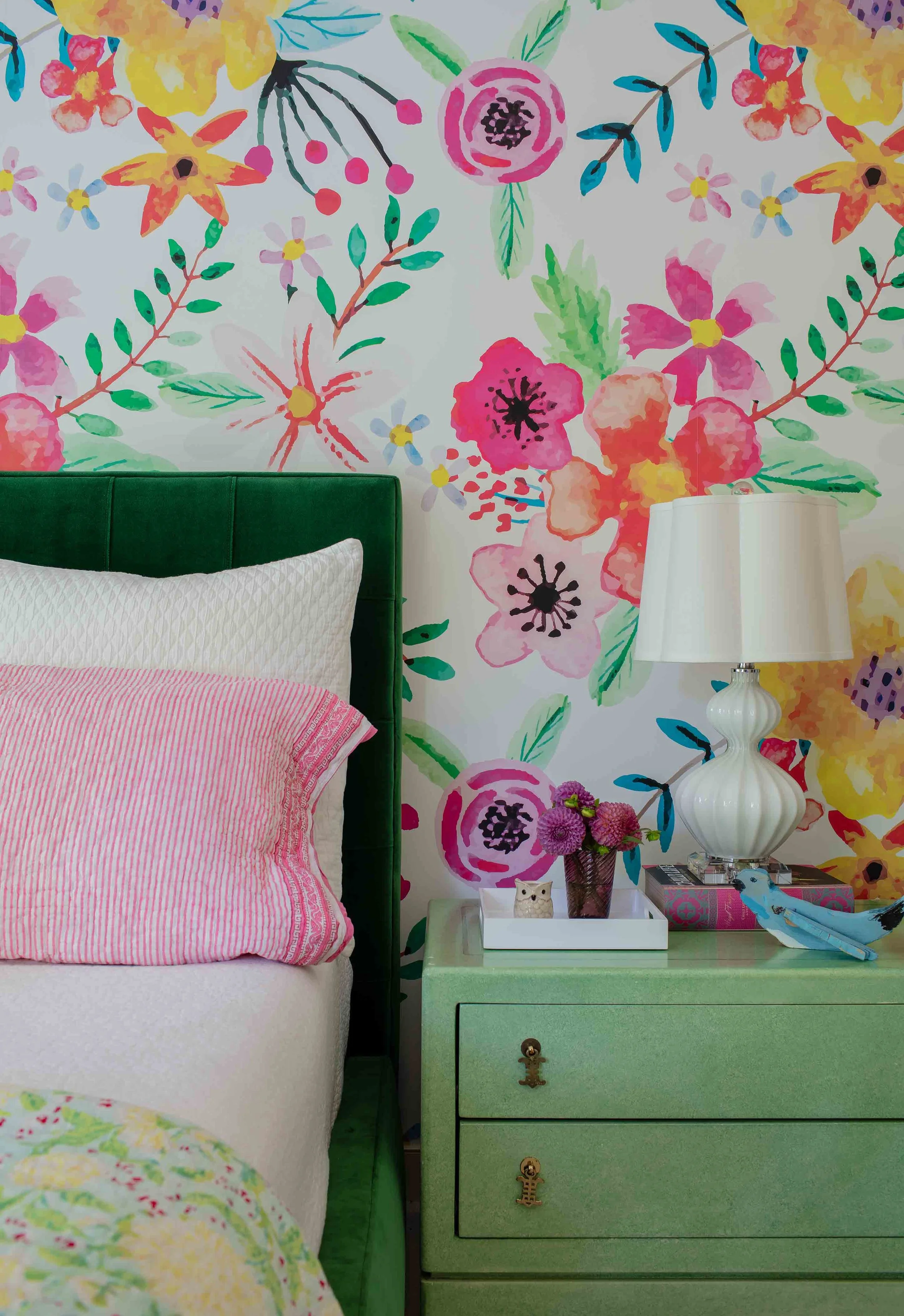 A bedroom with a green bedframe, pink and white bedding, a white nightstand with decorative items including a small owl figurine and pink flowers in a vase, a white table lamp, and a colorful floral wallpaper background.