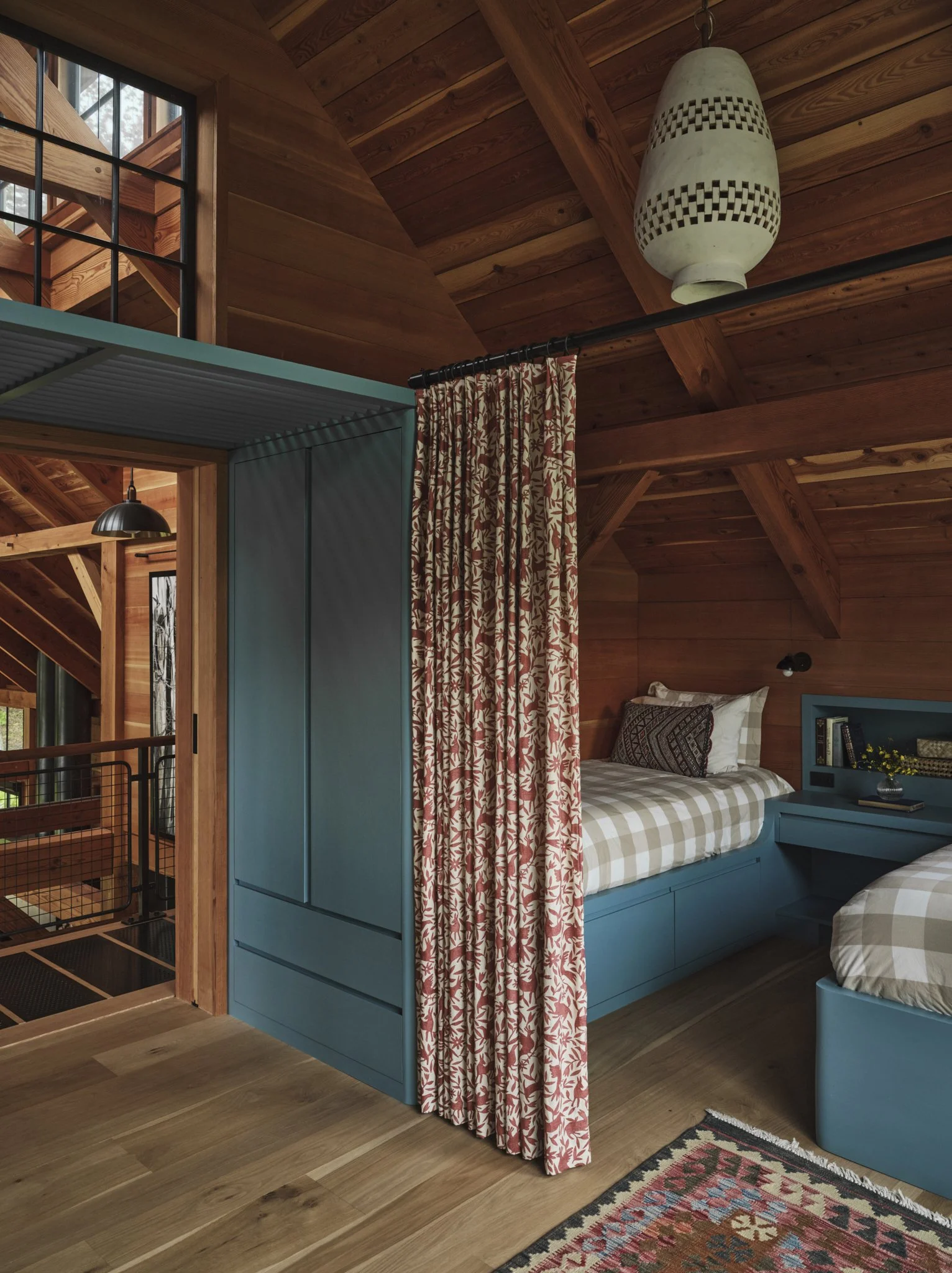 A cozy bedroom with wood-paneled walls and ceiling, featuring a bunk bed with floral and checkered bedding, blue furniture, and a patterned curtain.