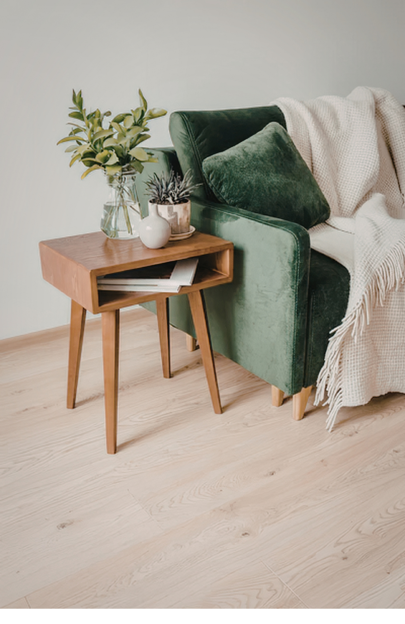 A cozy corner with a green velvet armchair, a wooden side table with plants, a cream throw blanket, and light hardwood flooring.