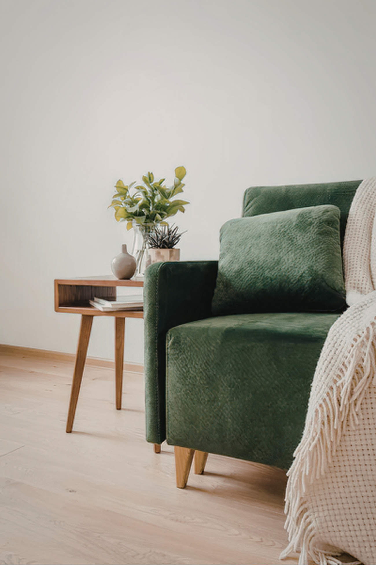 Living room with green velvet armchair, wooden side table with books, and a vase with yellow flowers on a minimalist background.