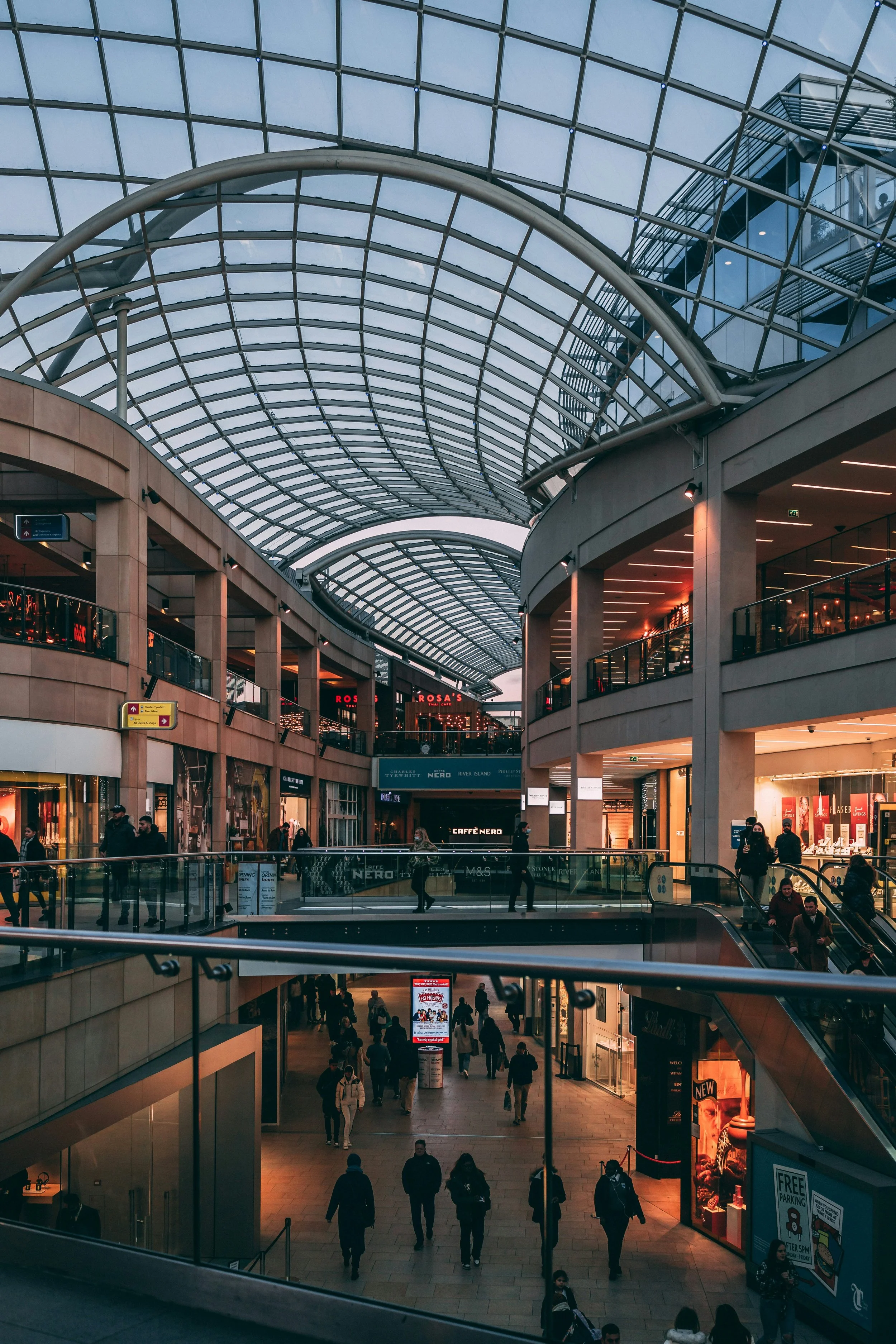 Intérieur moderne d'un centre commercial avec plusieurs étages, un toit en verre courbé et des boutiques.