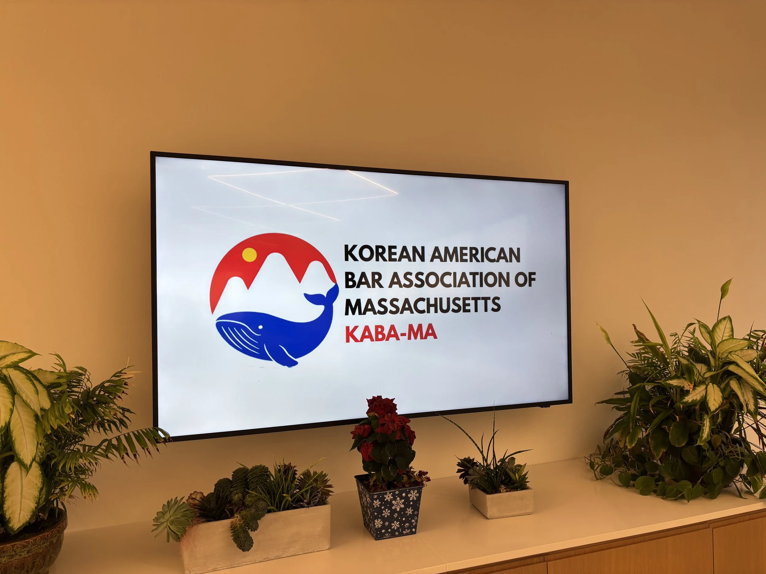 A screen displaying the logo and name of the Korean American Bar Association of Massachusetts, surrounded by potted plants on a wooden surface.