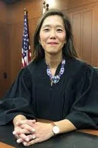 A woman in a judicial robe sitting at a judge's bench with an American flag in the background.
