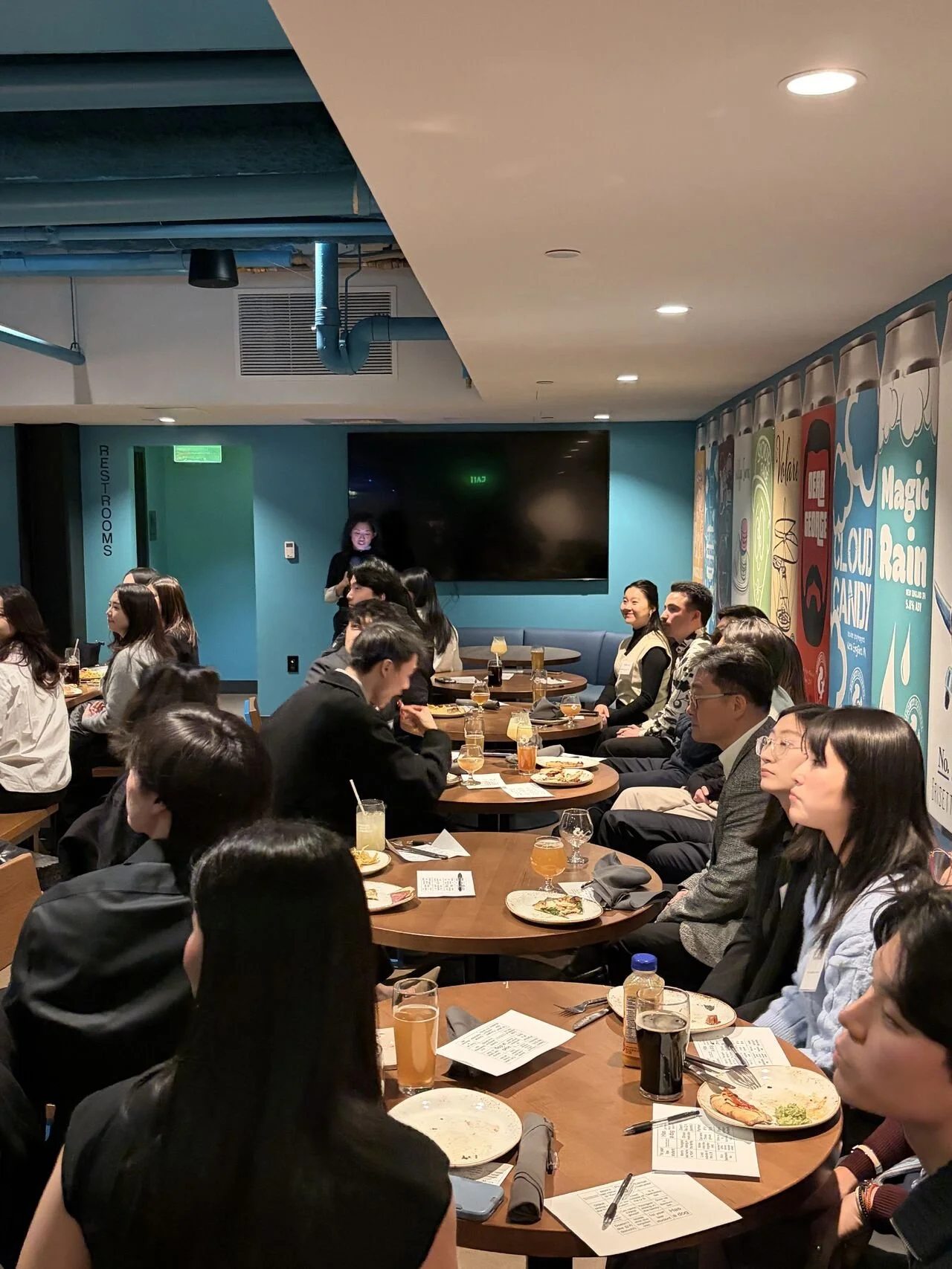 People seated at round tables in a restaurant, attending an event or gathering, with one woman standing near a large black screen on the teal-colored wall and colorful artwork depicting various comic-style images and text.