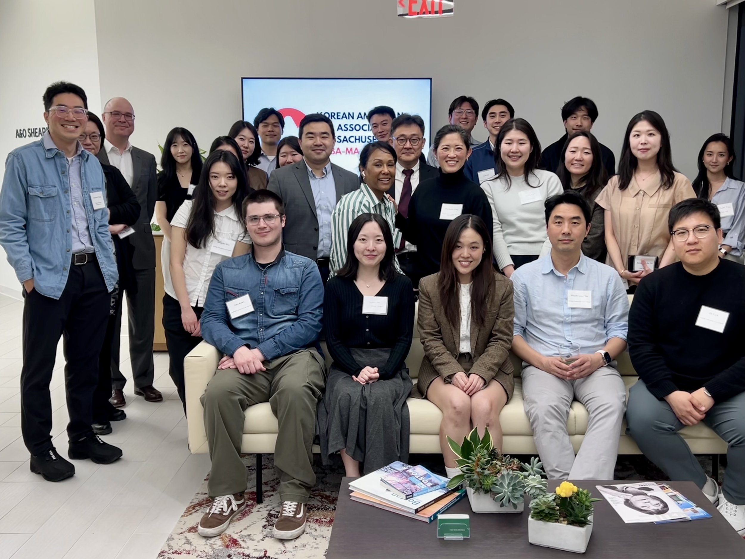 A large group of people, diverse in age and ethnicity, gathered in a room for a group photo. They are smiling and some are wearing name tags. Behind them, a sign on the wall reads 'Korean American Association of Massachusetts' and there's a screen wi