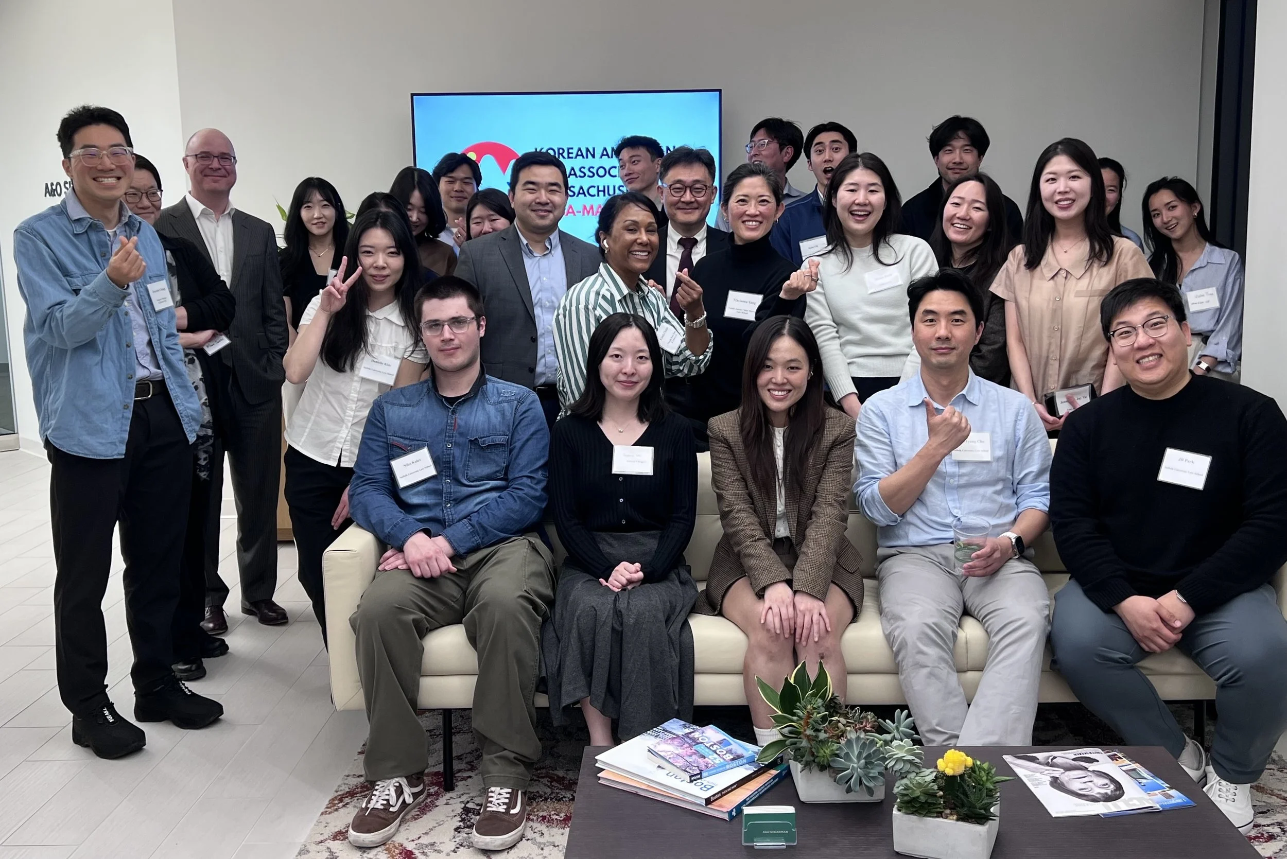 Group of diverse people smiling and making hand gestures at a professional event in a room with a large screen displaying 'Korean Americans Association' and a table with books and plants in the foreground.