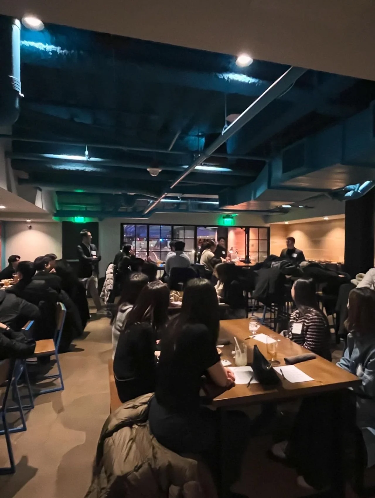 A crowded social event or conference in a restaurant or cafe setting with many people seated at tables, some standing and speaking, in a dimly lit room with large windows and modern decor.