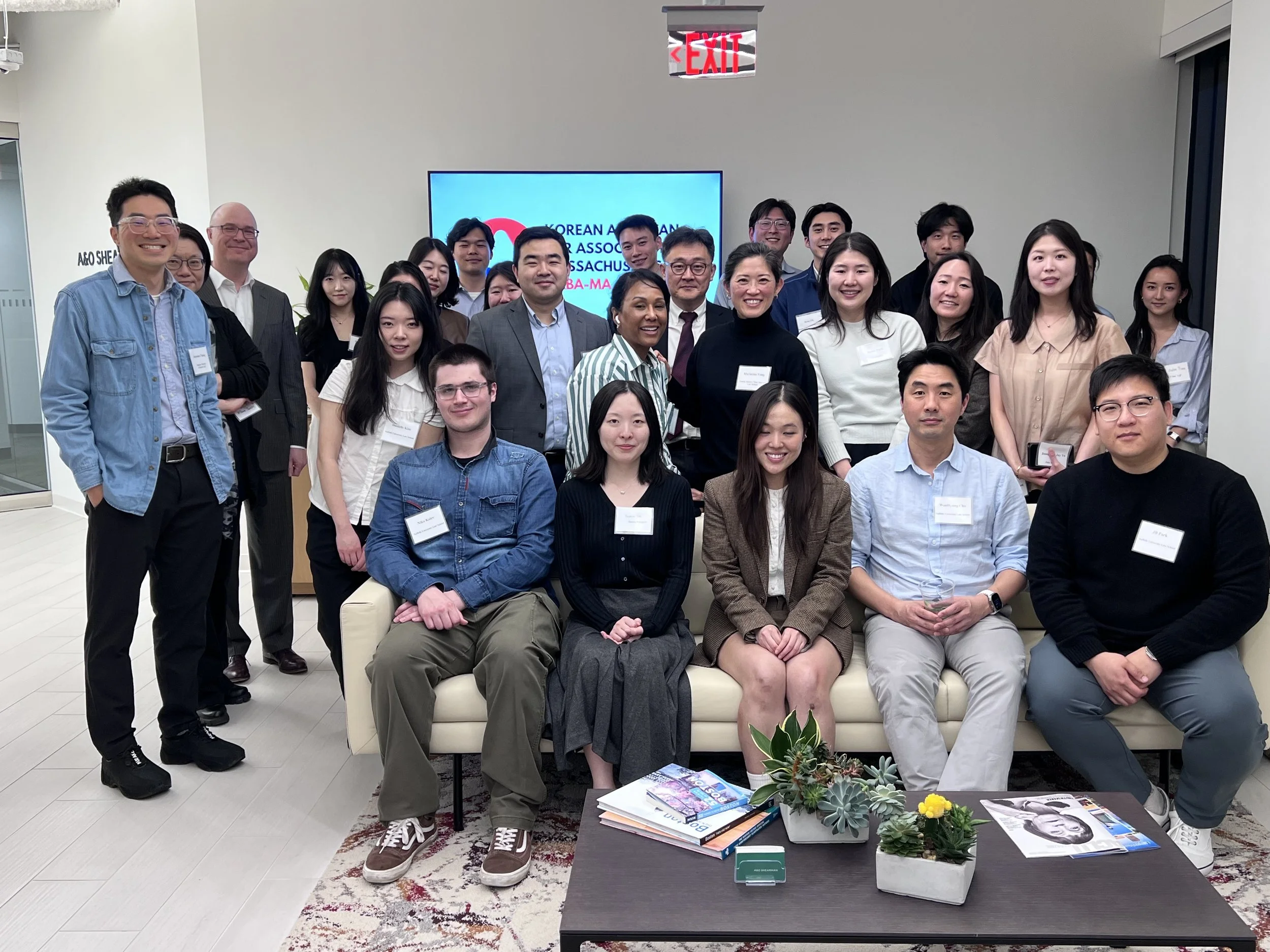 Group of diverse people in an office, smiling, standing and sitting, with a blue screen in the background displaying a logo and text. There are plants and magazines on a table in the foreground.