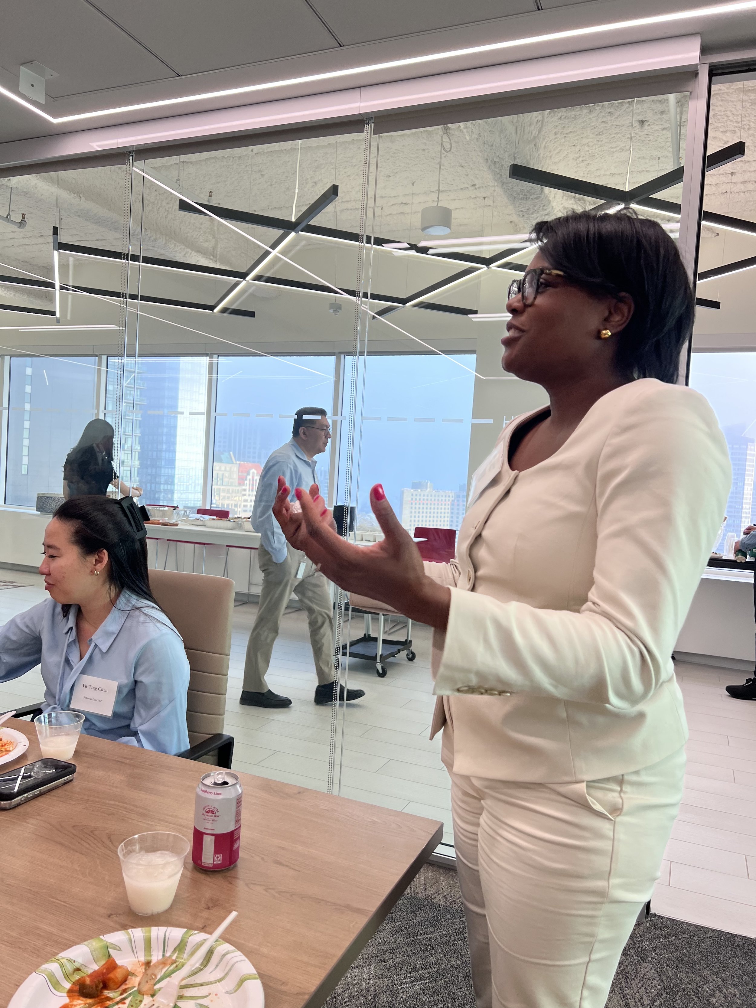 A professional meeting or conference room with modern geometric ceiling lighting and large windows, showing a woman with short black hair, glasses, and gold earrings talking. Other people are seated at a table with plates and drinks, and a man in a l
