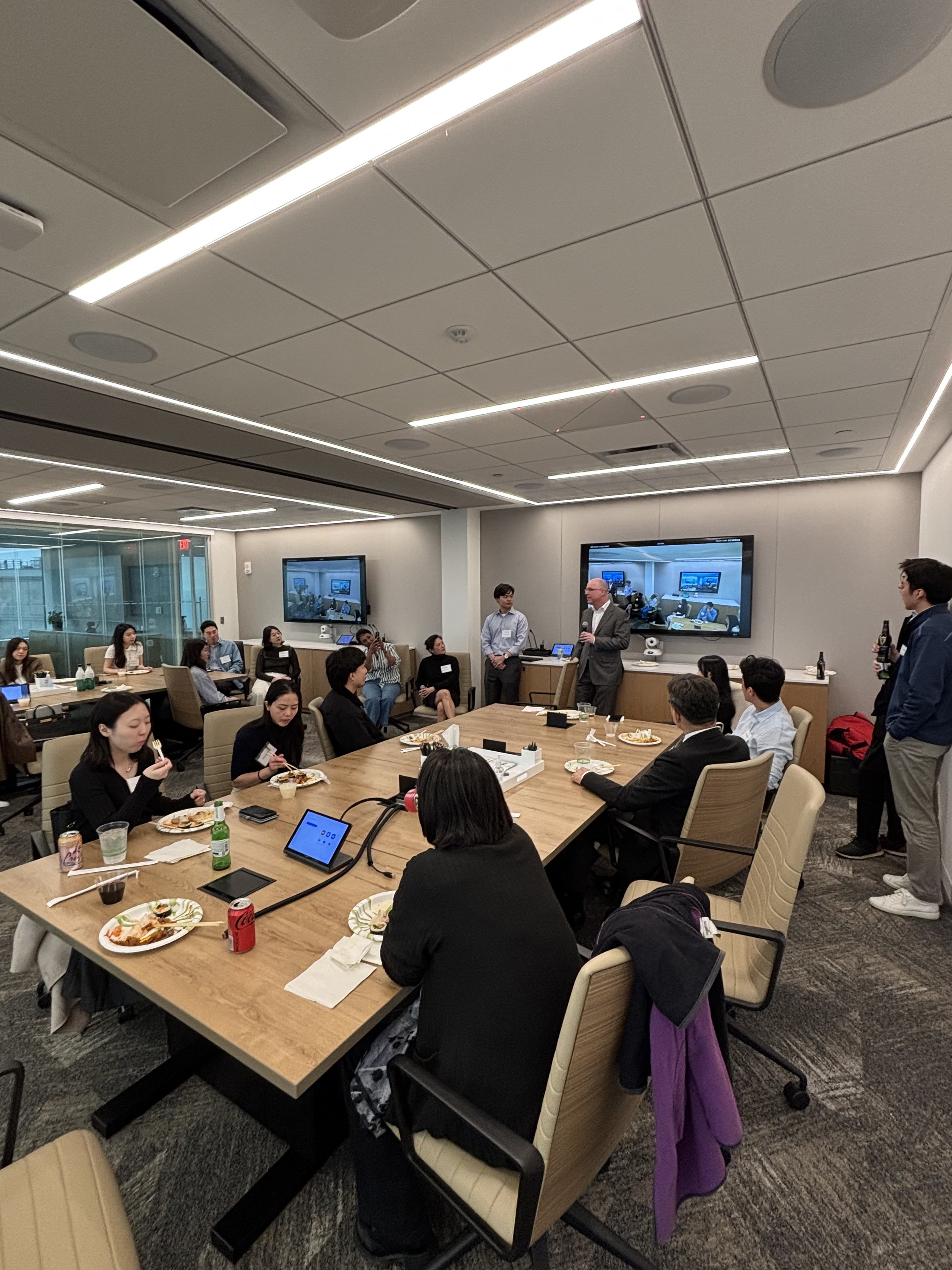 A conference or meeting room with people sitting around tables, some eating and drinking, while speakers stand near a large screen and a TV monitor, engaging with the audience.