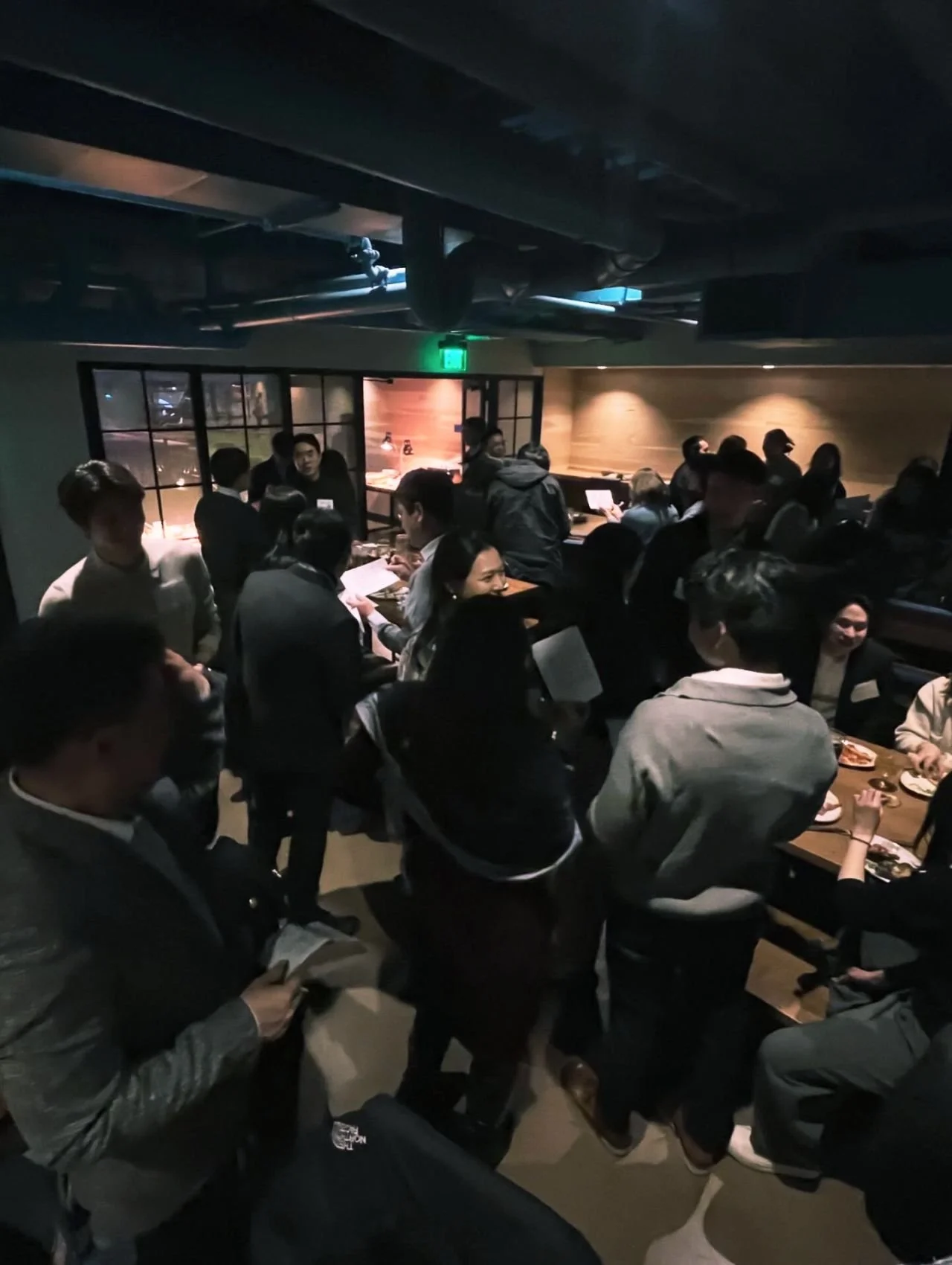 Crowded restaurant or cafe with people standing and sitting, some engaged in conversation or using electronic devices, warm lighting, and a modern interior design.