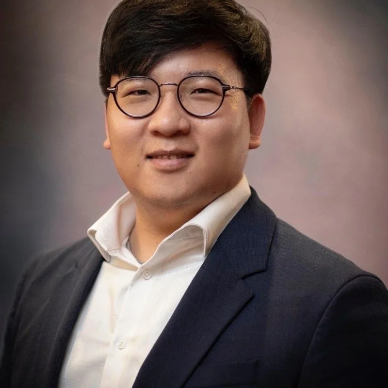 Portrait of a young man with glasses wearing a white shirt and dark blazer, smiling against a blurred background.