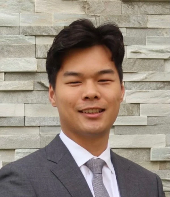 A young man dressed in a gray suit with a white shirt and gray tie, smiling in front of a stone brick wall.