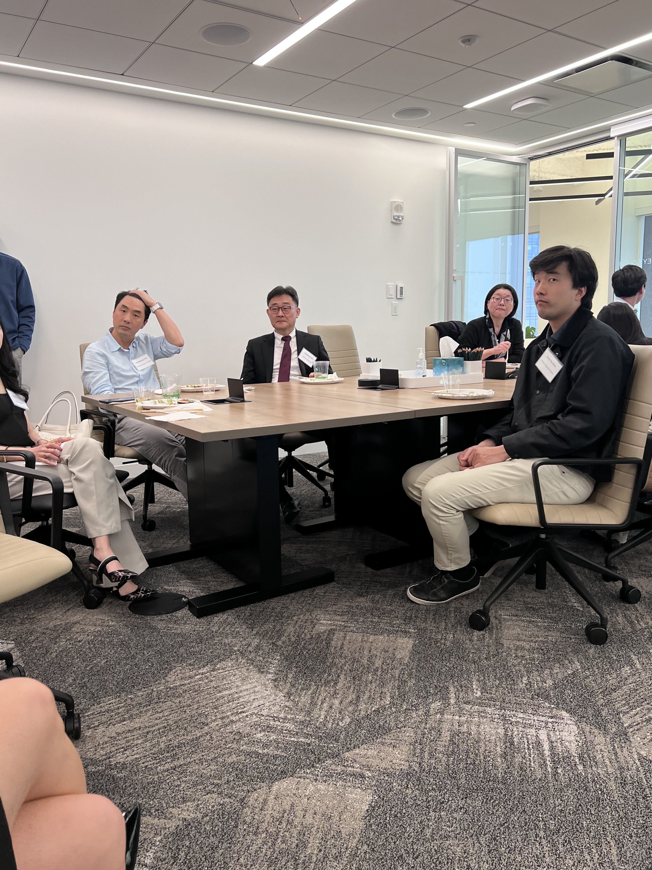 People seated around a conference table in a modern office meeting room. Some appear to be listening, while others look contemplative. There are plates, drinks, and office supplies on the table, with a large window showing the outside.