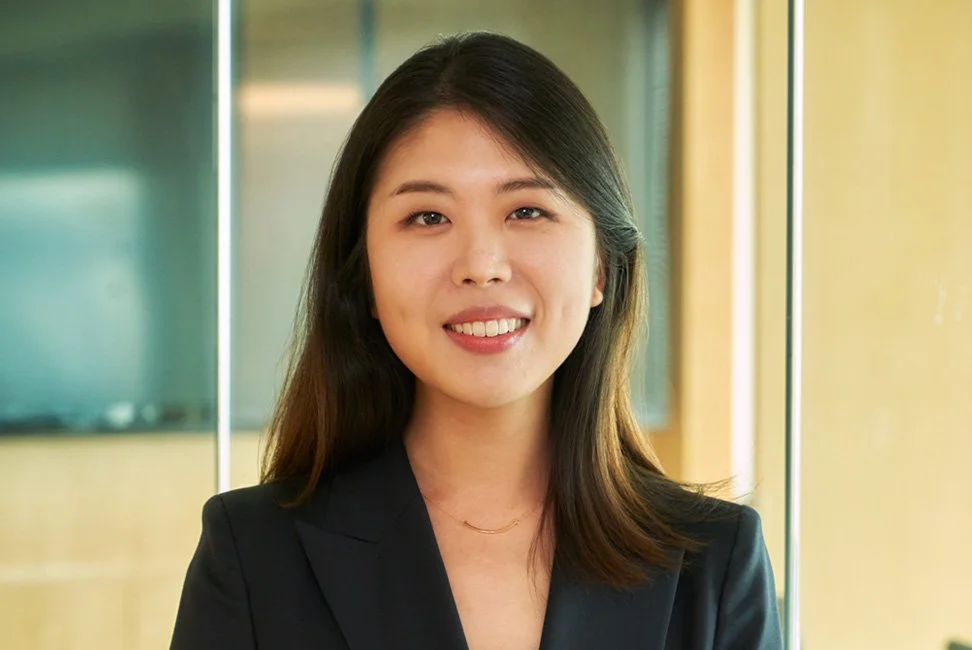 A young woman with shoulder-length dark hair, smiling, wearing a black blazer, standing in an office with glass walls.