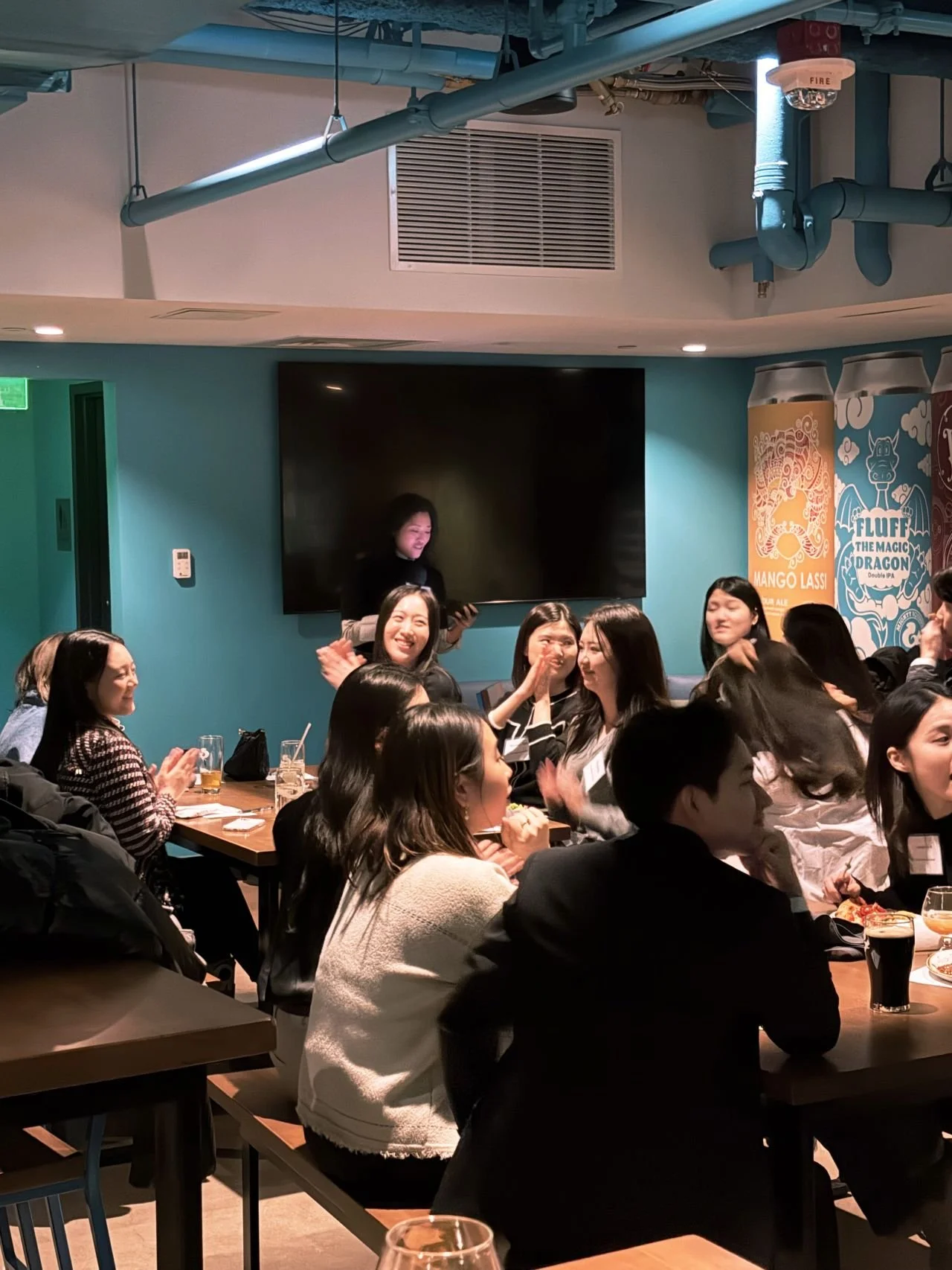 Group of women and men gathered at long tables in a restaurant, appearing to celebrate or enjoy an event, with some smiling, talking, and clapping. There are drinks, plates of food, and a large screen on the wall behind them. The decor includes a mural with beer cans and a dragon cartoon, and the ceiling has blue pipes.