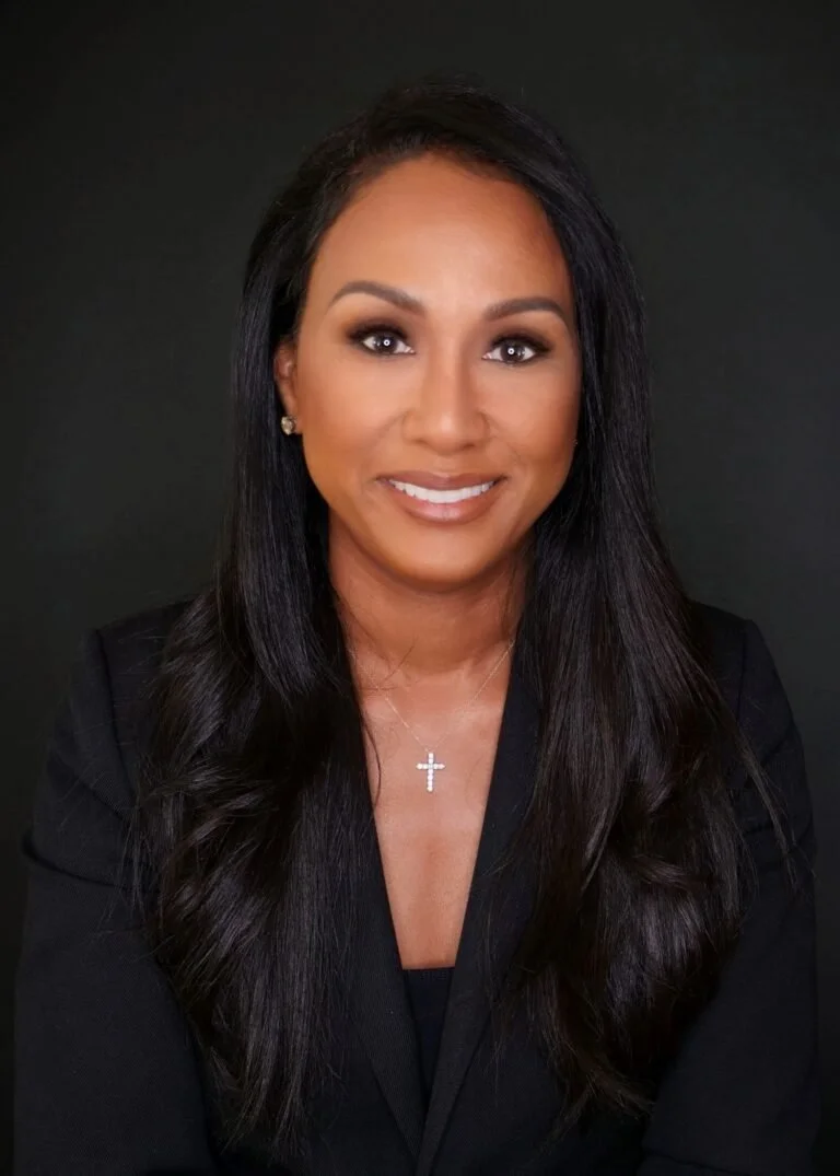 A woman with long dark hair, wearing a black blazer, small earrings, a cross necklace, and smiling against a dark background.