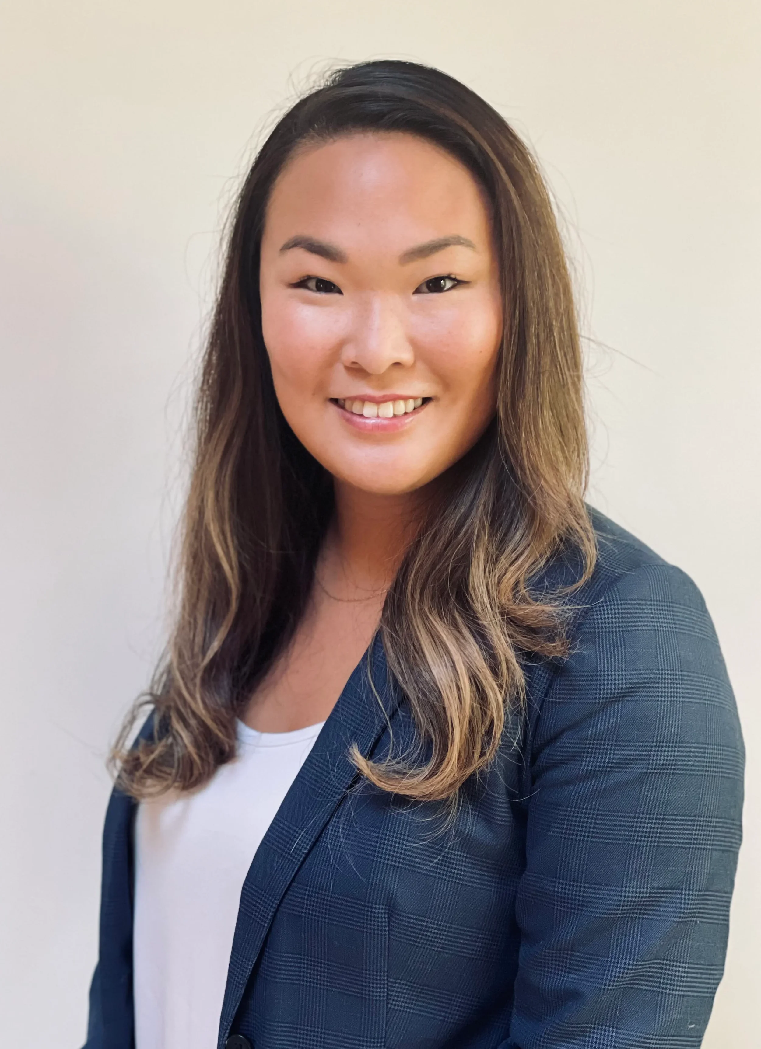 A smiling woman with medium skin tone, long wavy hair, and wearing a blue checkered blazer over a white top, standing against a plain light-colored background.