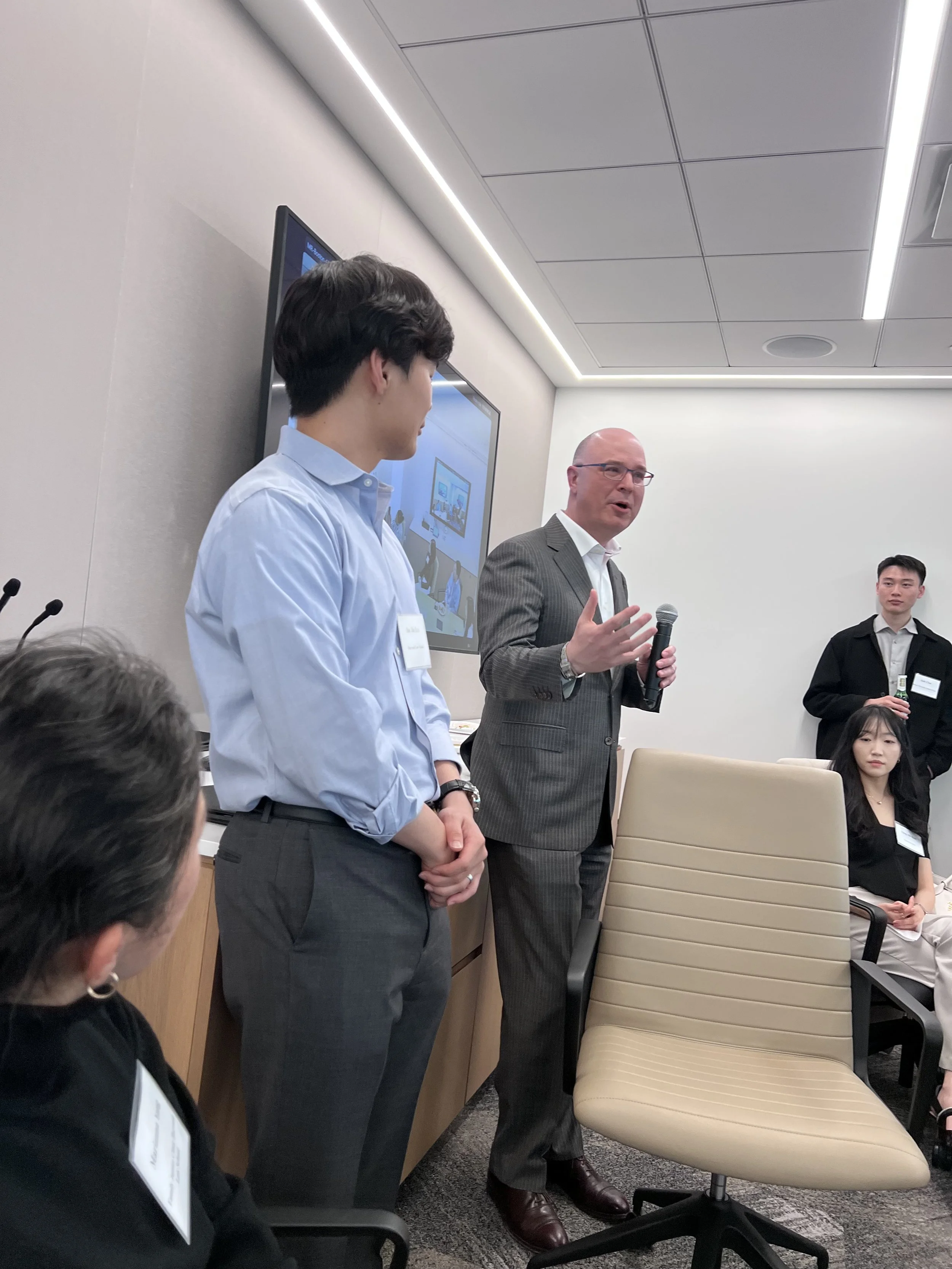 A man in a suit and glasses speaking into a microphone while three other people listen. One person is dressed in a white shirt, another in a black blazer, and a woman in a black outfit is seated. There is a large screen in the background showing the 