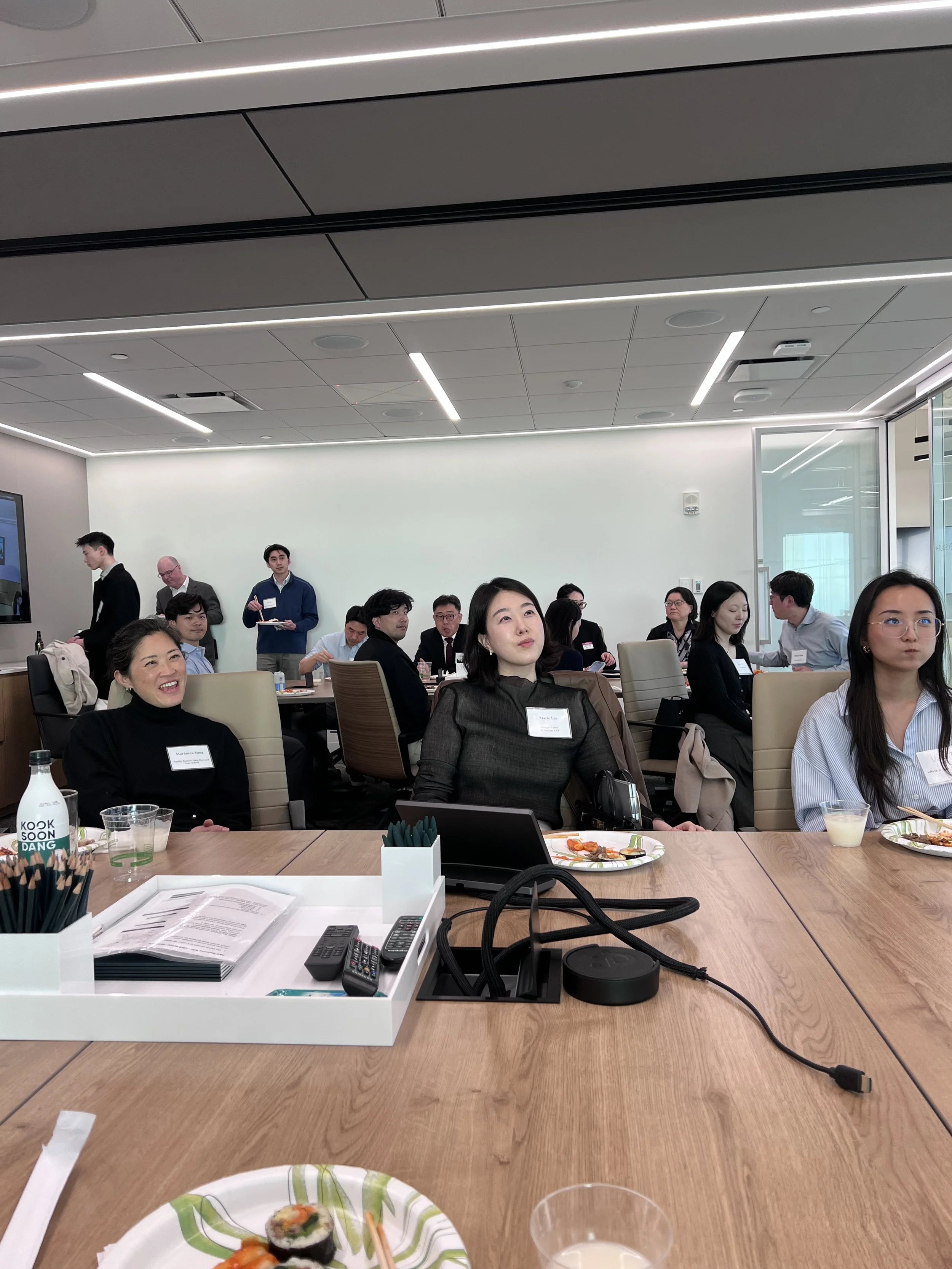 A diverse group of people attending a professional meeting or conference in a modern, well-lit conference room. Attendees are sitting around a large table, some are eating, and others are listening attentively. There are plates of food, drinks, and c