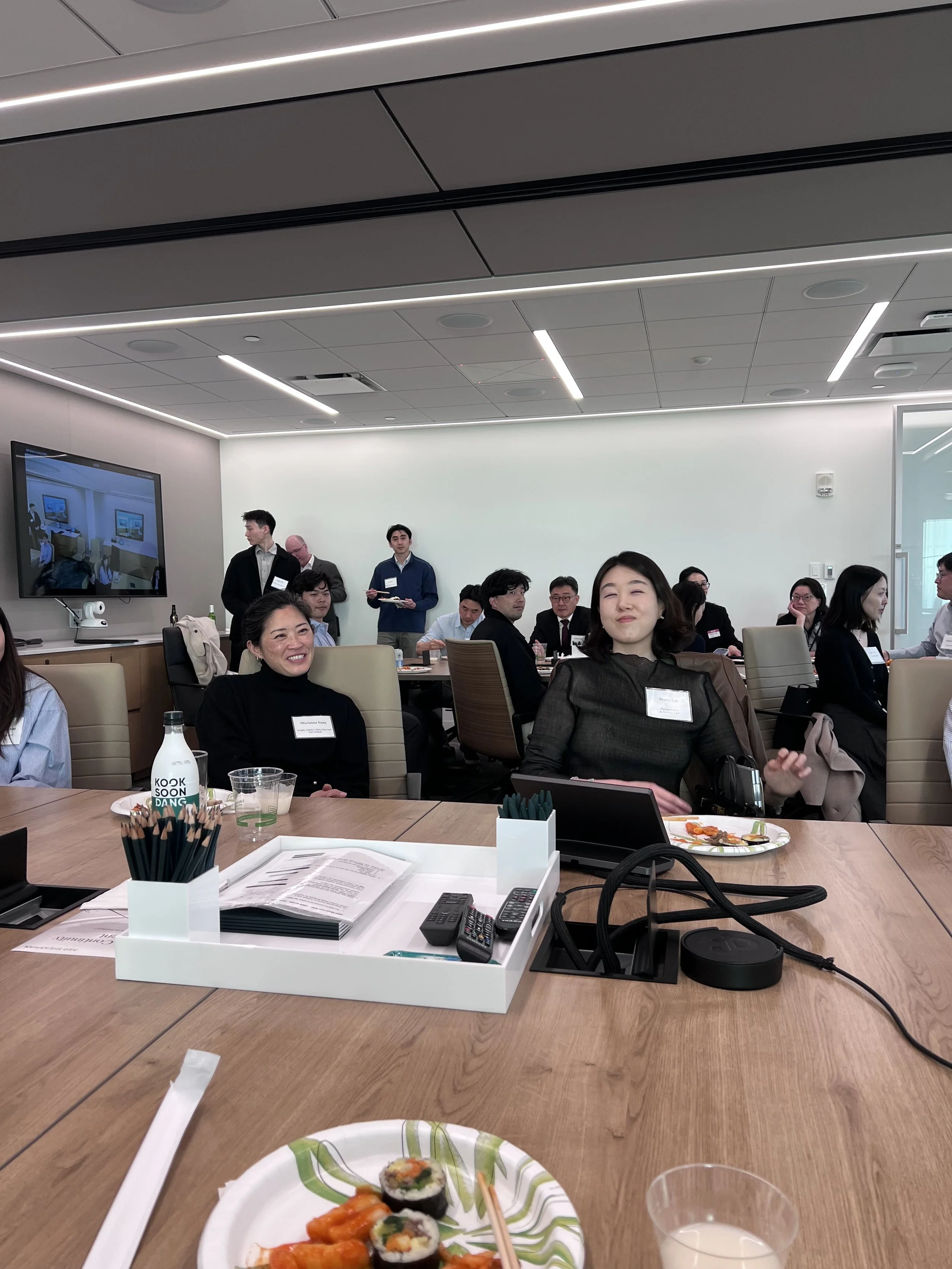 A meeting room with people sitting around a large wooden table, some smiling and others focused. In the foreground, there's a plate of sushi with chopsticks, a cup with a beverage, and a dish of food. The background includes a television screen on th