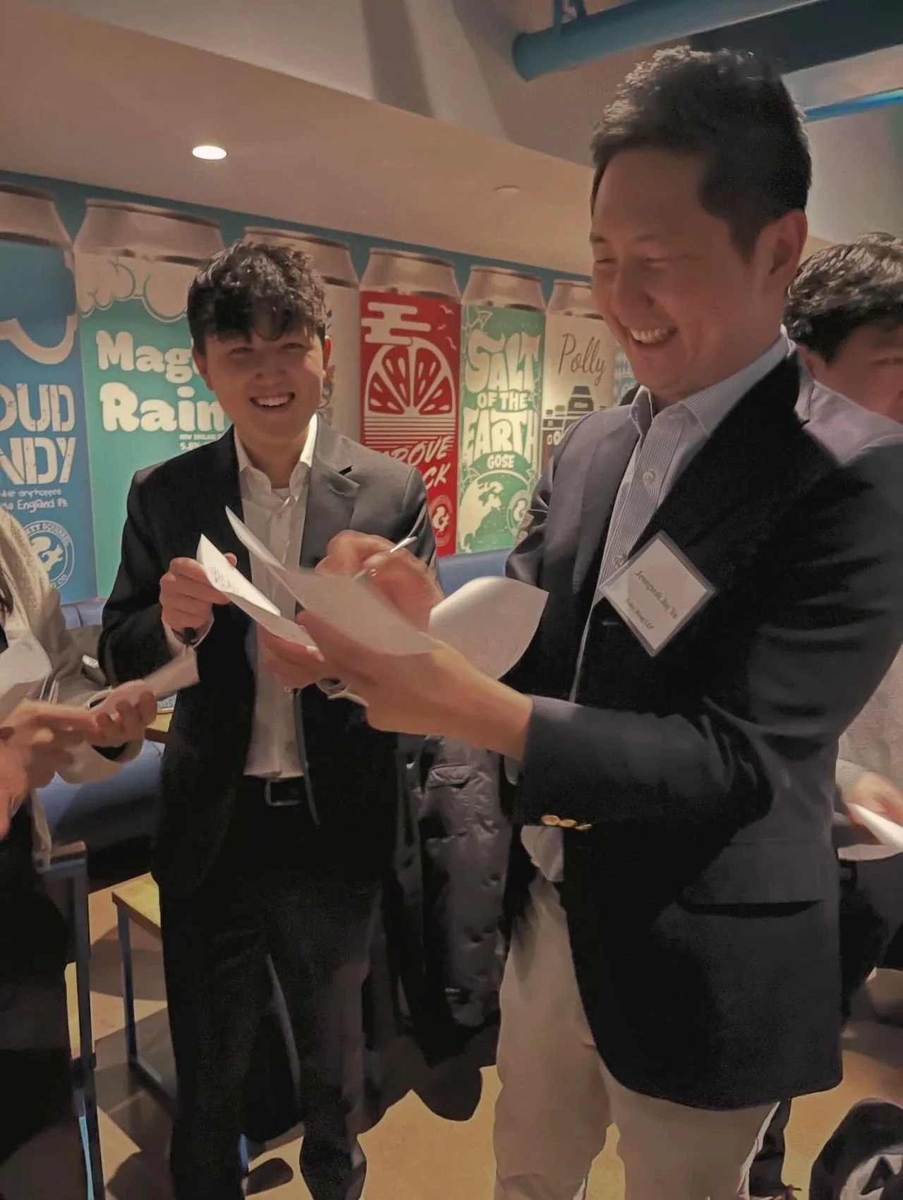 Two men in suits smiling and exchanging papers at a social event, with colorful posters on the wall behind them.