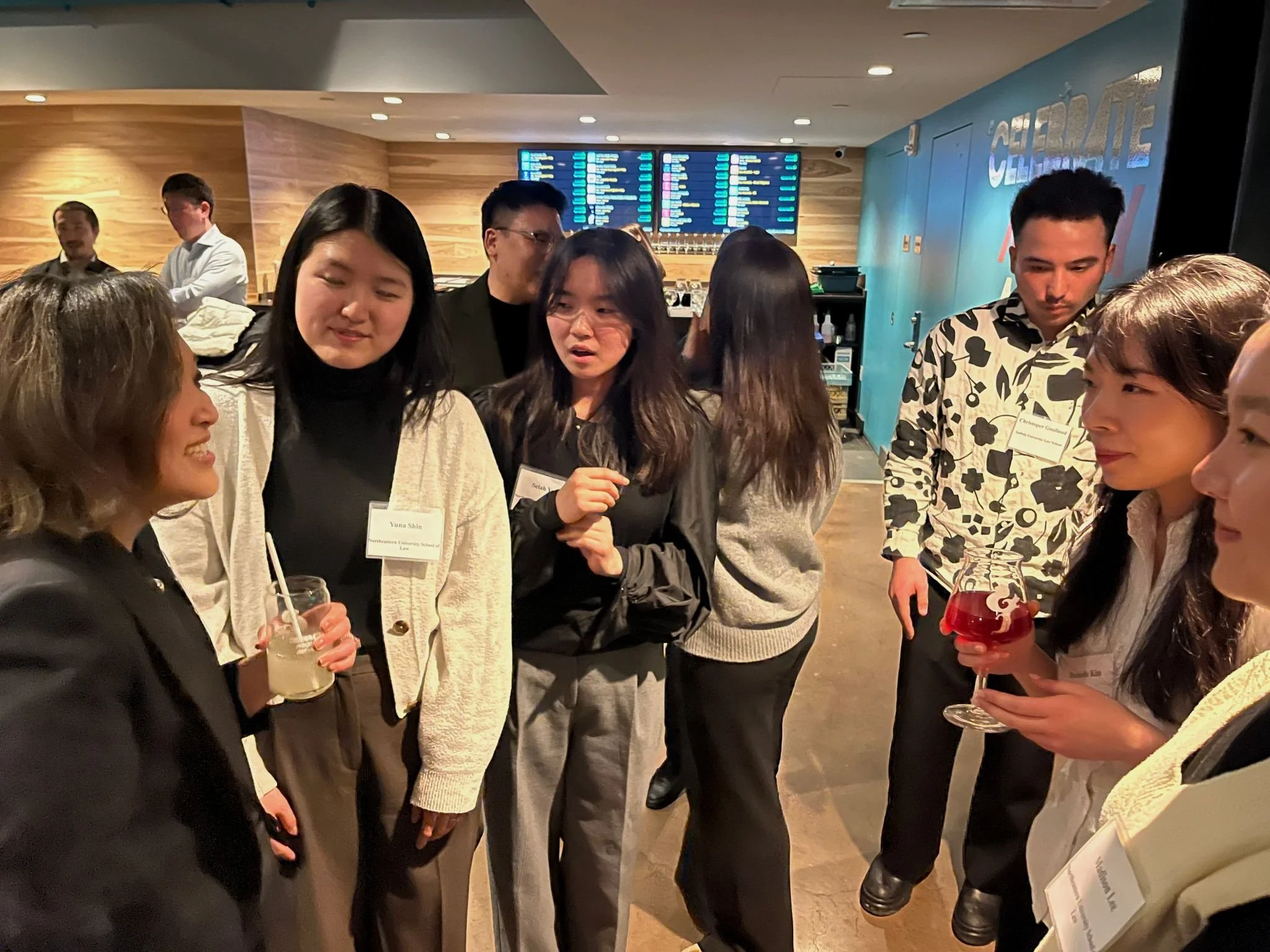 Group of young adults socializing at a busy indoor venue, holding drinks, with an electronic flight information display in the background.