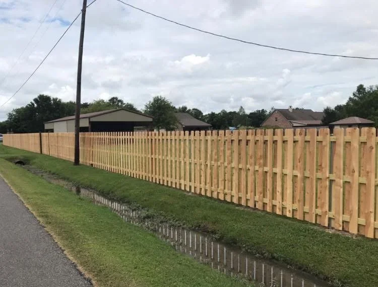 A wooden privacy fence runs alongside a grassy area next to a street with a drainage ditch. There are houses and trees in the background under a cloudy sky.