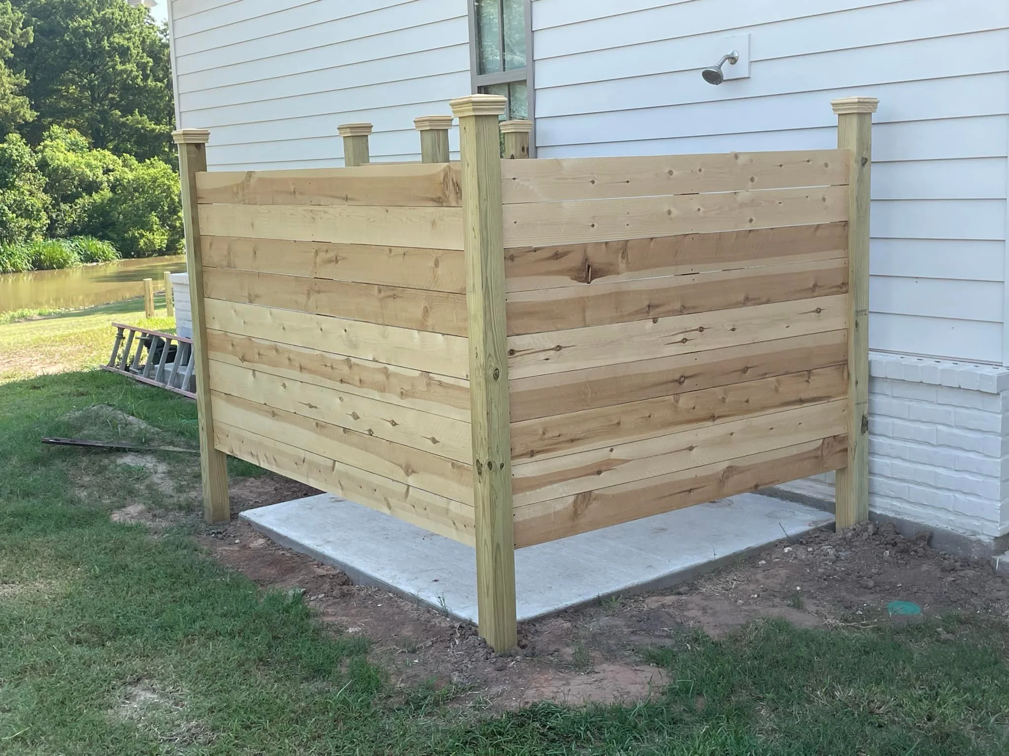A wooden privacy fence installed around a small concrete slab outside a white house.