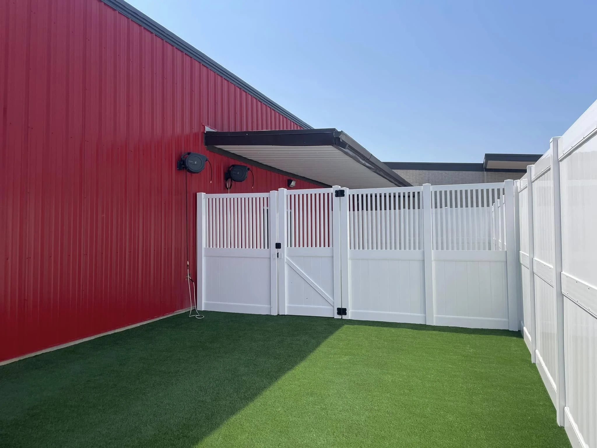 Small backyard with artificial grass, red and white fencing, and a red building wall with two black outdoor lights under a small overhang.