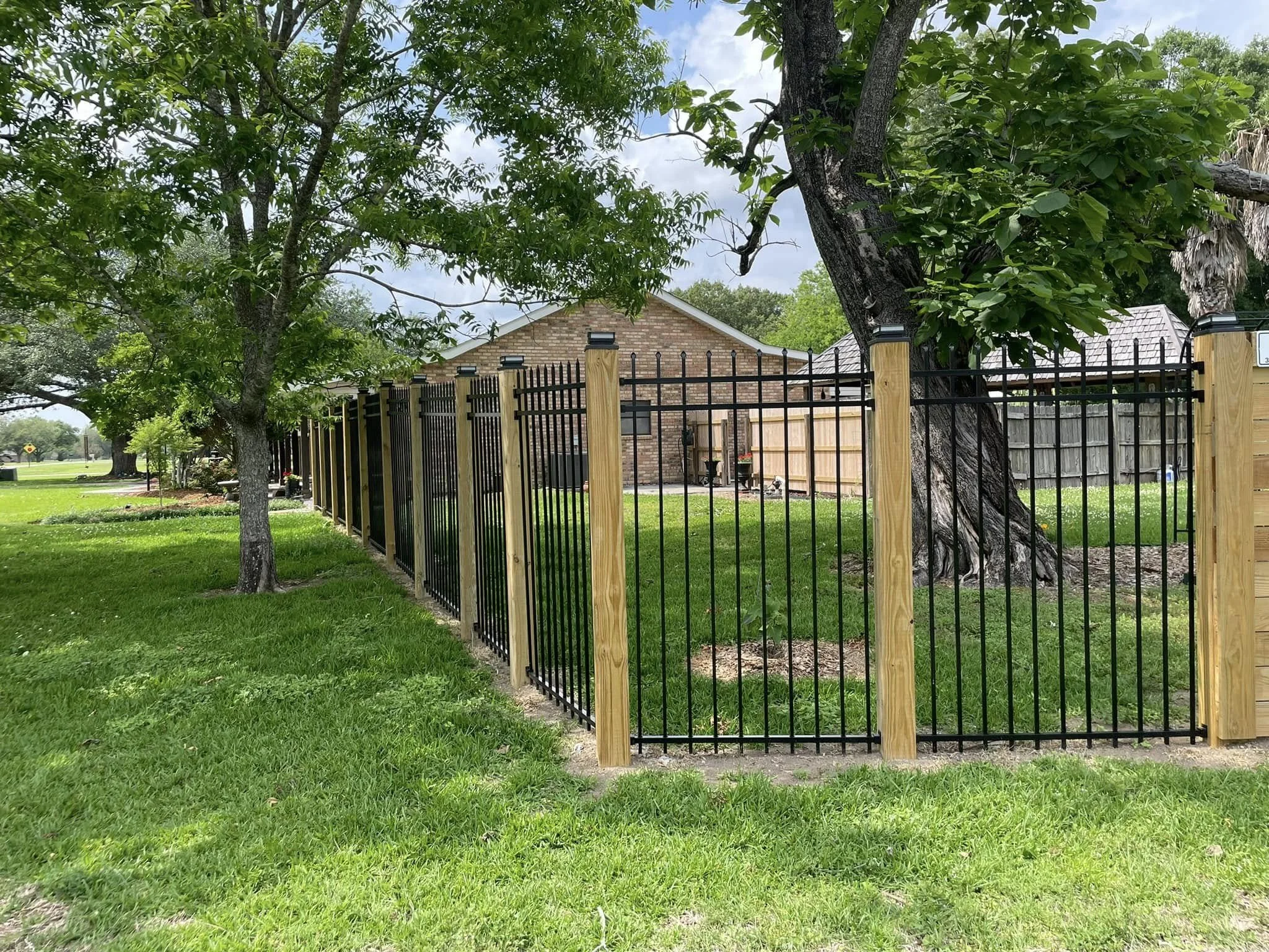 A fenced backyard with green grass, trees, and a brick house in the background.