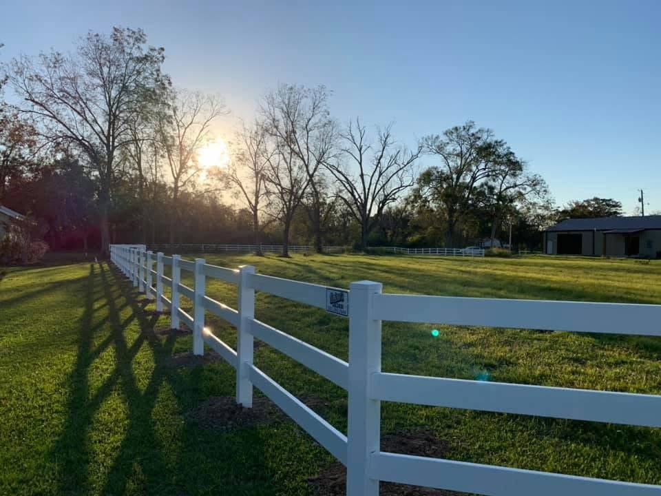 A white fence running along a grassy field with trees and a building in the background, sun setting in the sky.