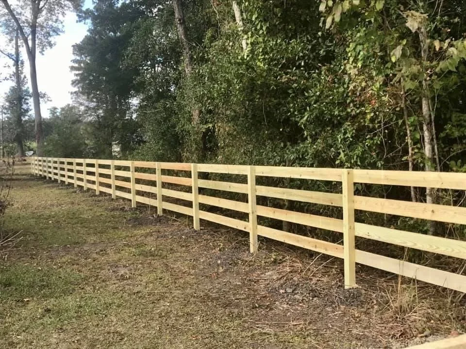 New wooden fence along a grassy and dirt area beside a dense tree line.