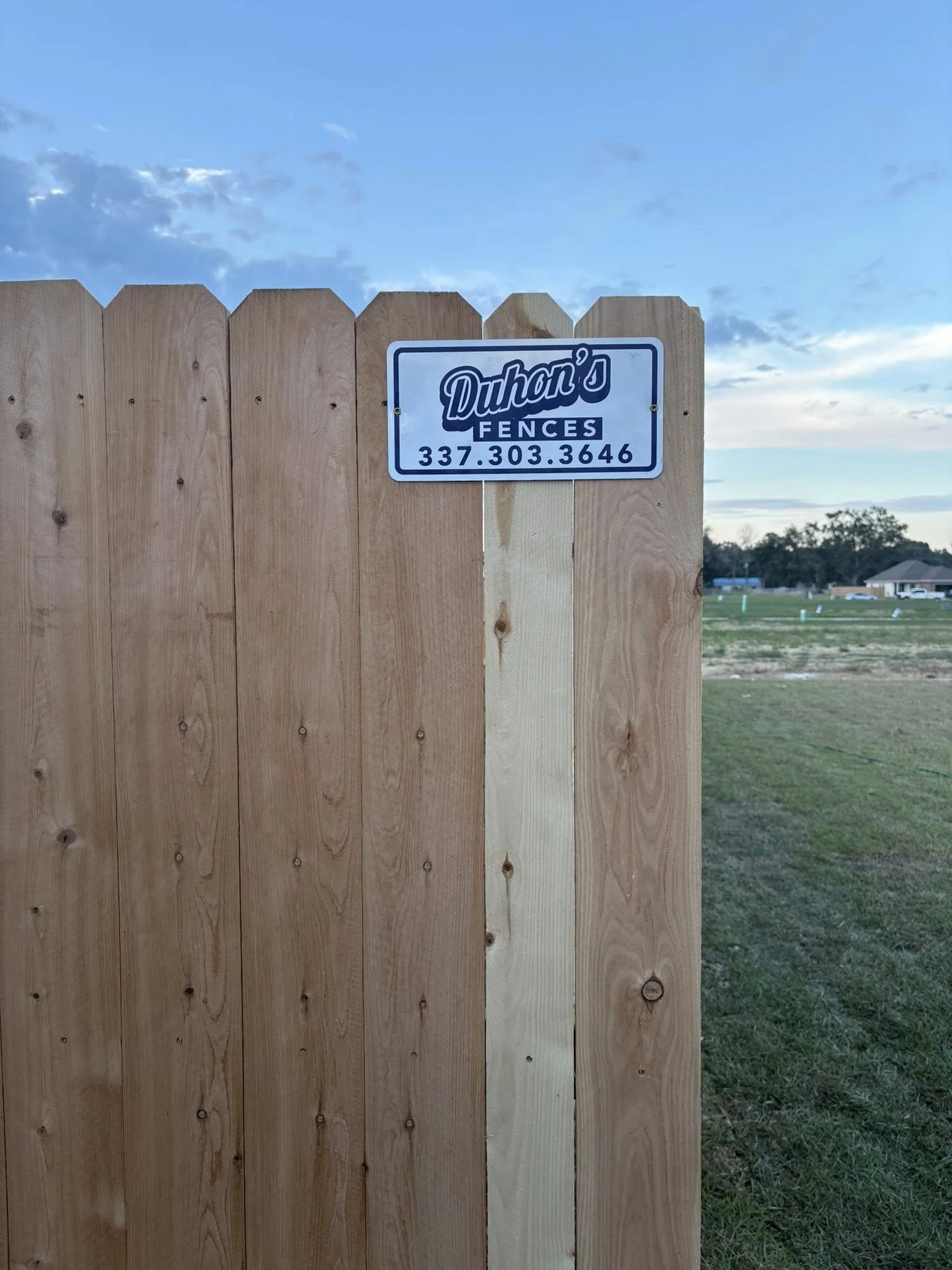 A newly installed wooden fence with a sign that reads 'Duhon's Fences' and a phone number, set in a grassy field with a few houses and trees in the background under a partly cloudy sky at dusk.