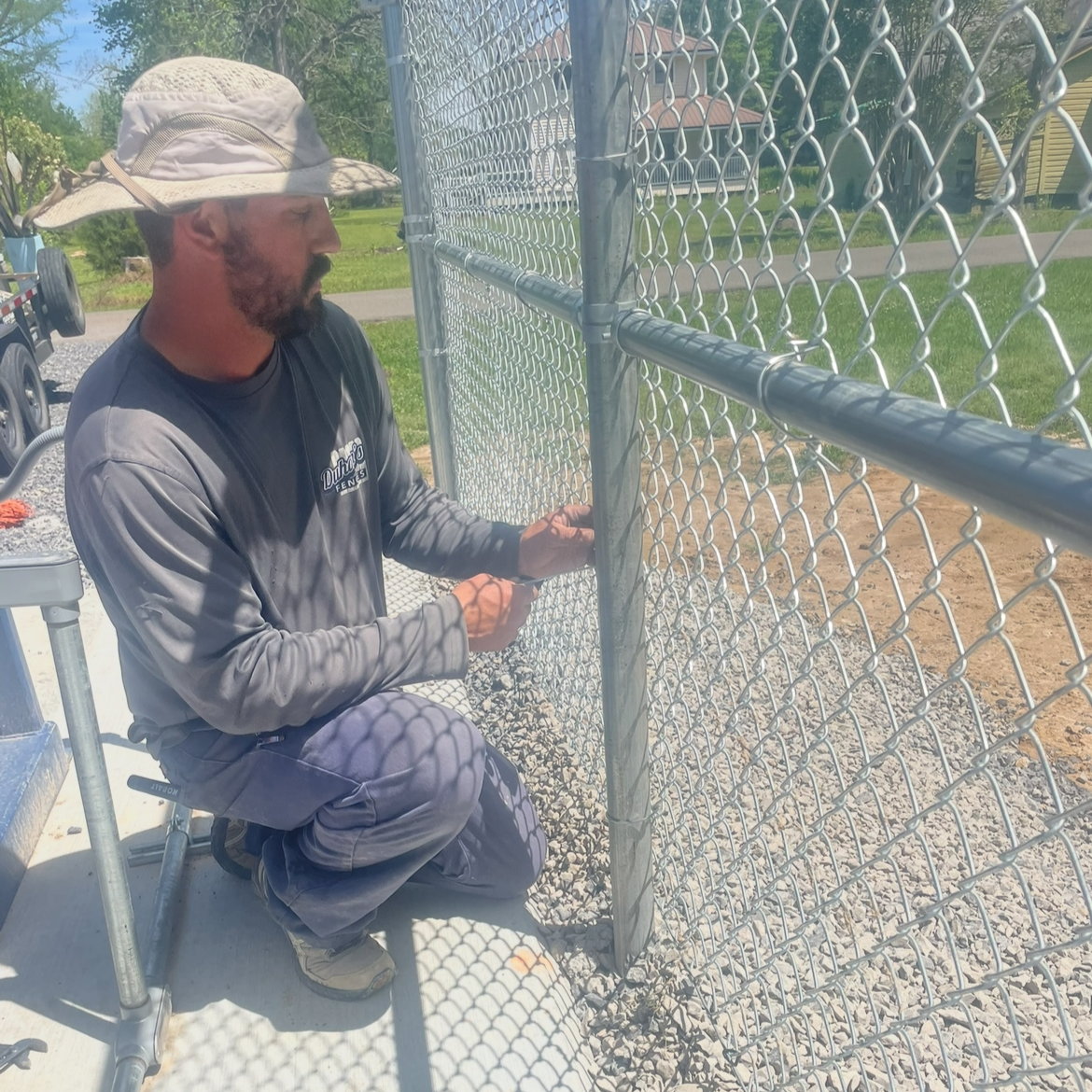 A man kneeling on a raised platform, installing a chain-link fence outdoors on a sunny day.