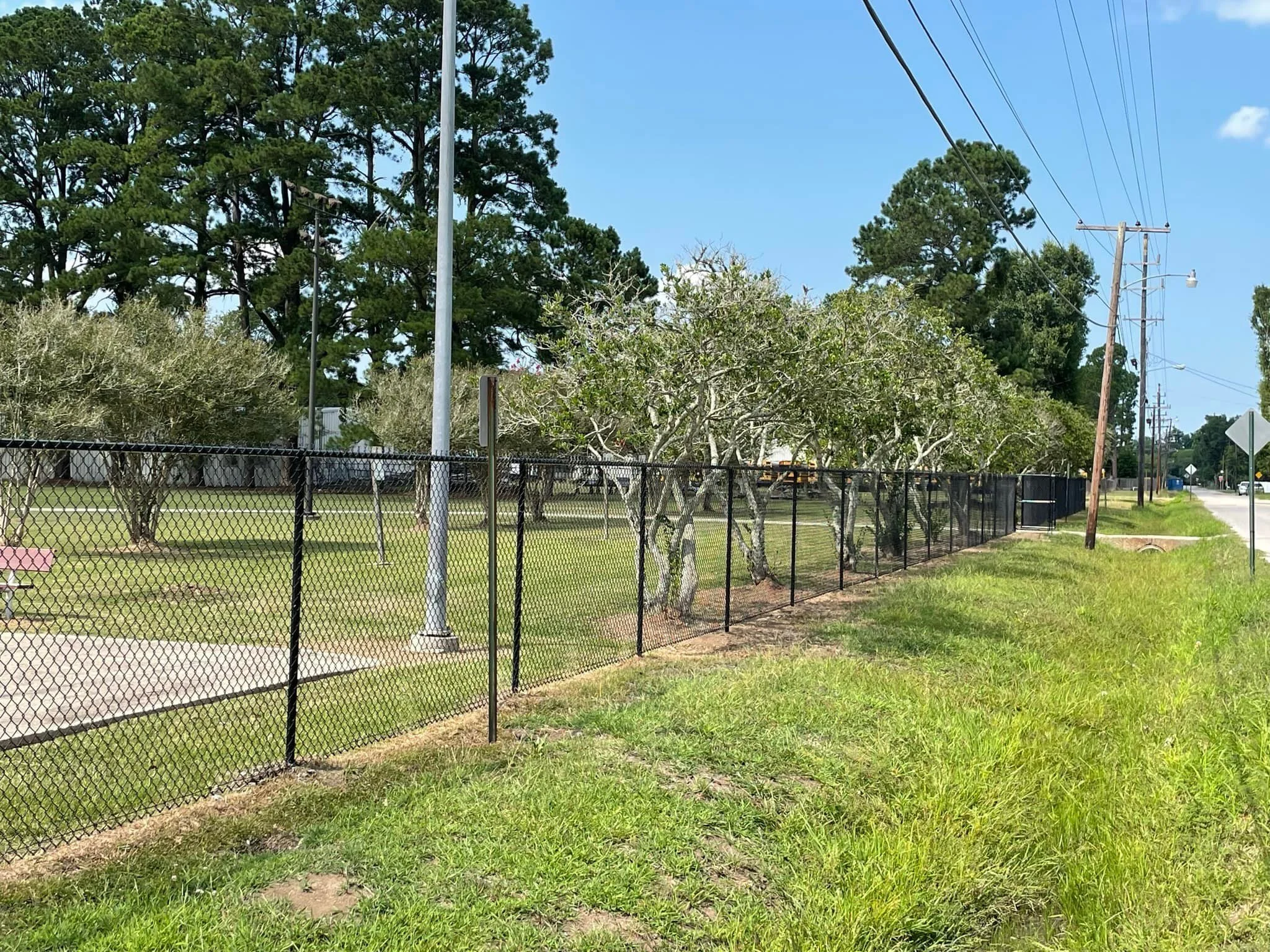 A black metal fence encloses a grassy park area with trees, with utility poles and power lines along the street beside it. The sky is blue with a few clouds.
