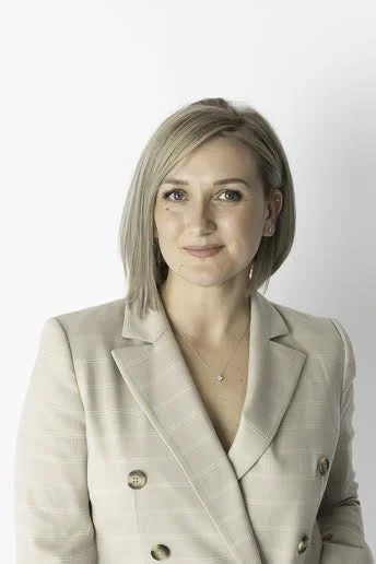 A woman with short blonde hair, wearing a light-colored blazer, standing against a plain white background.