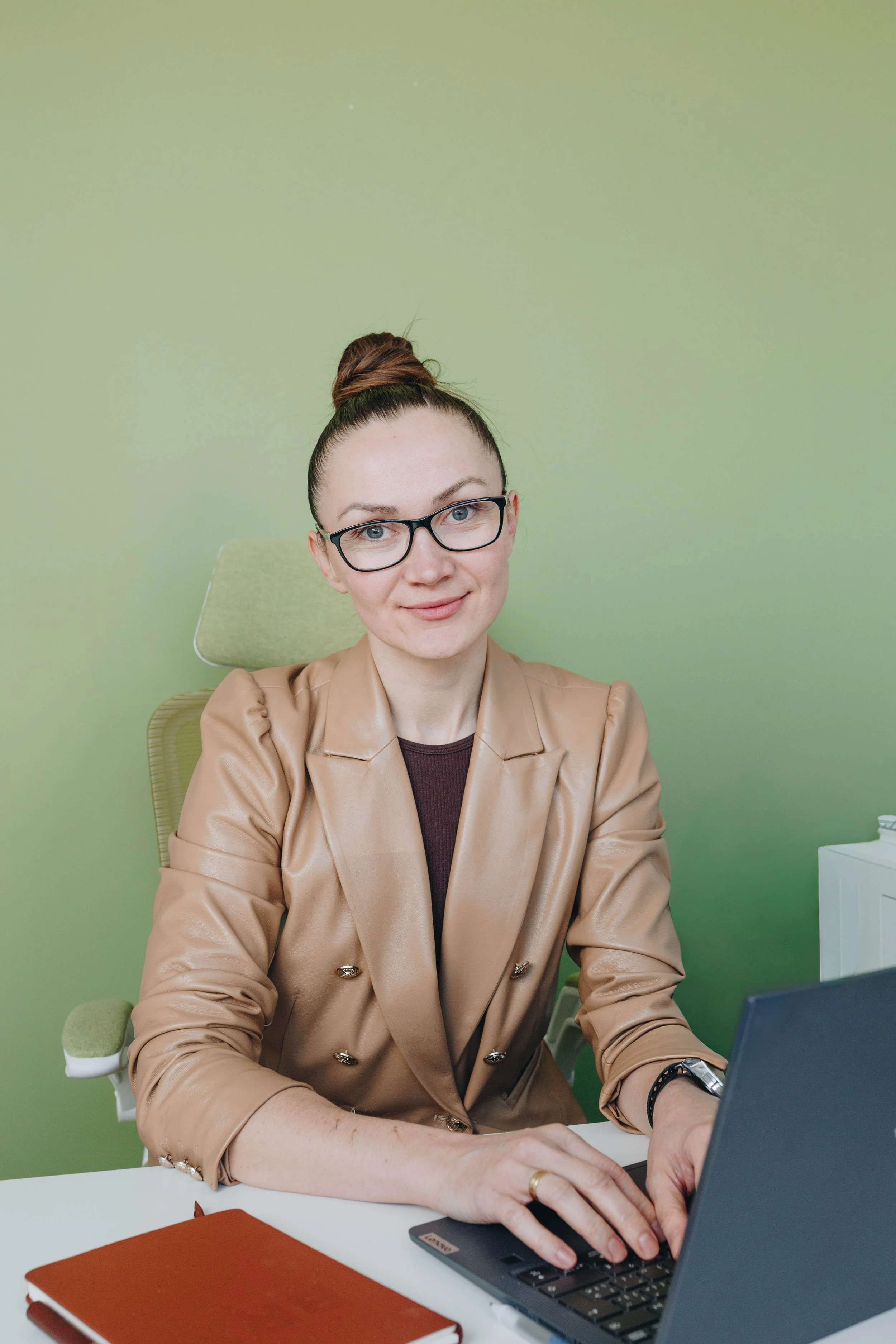 A woman with glasses and a bun hairstyle working on a laptop at a desk in front of a light green wall.