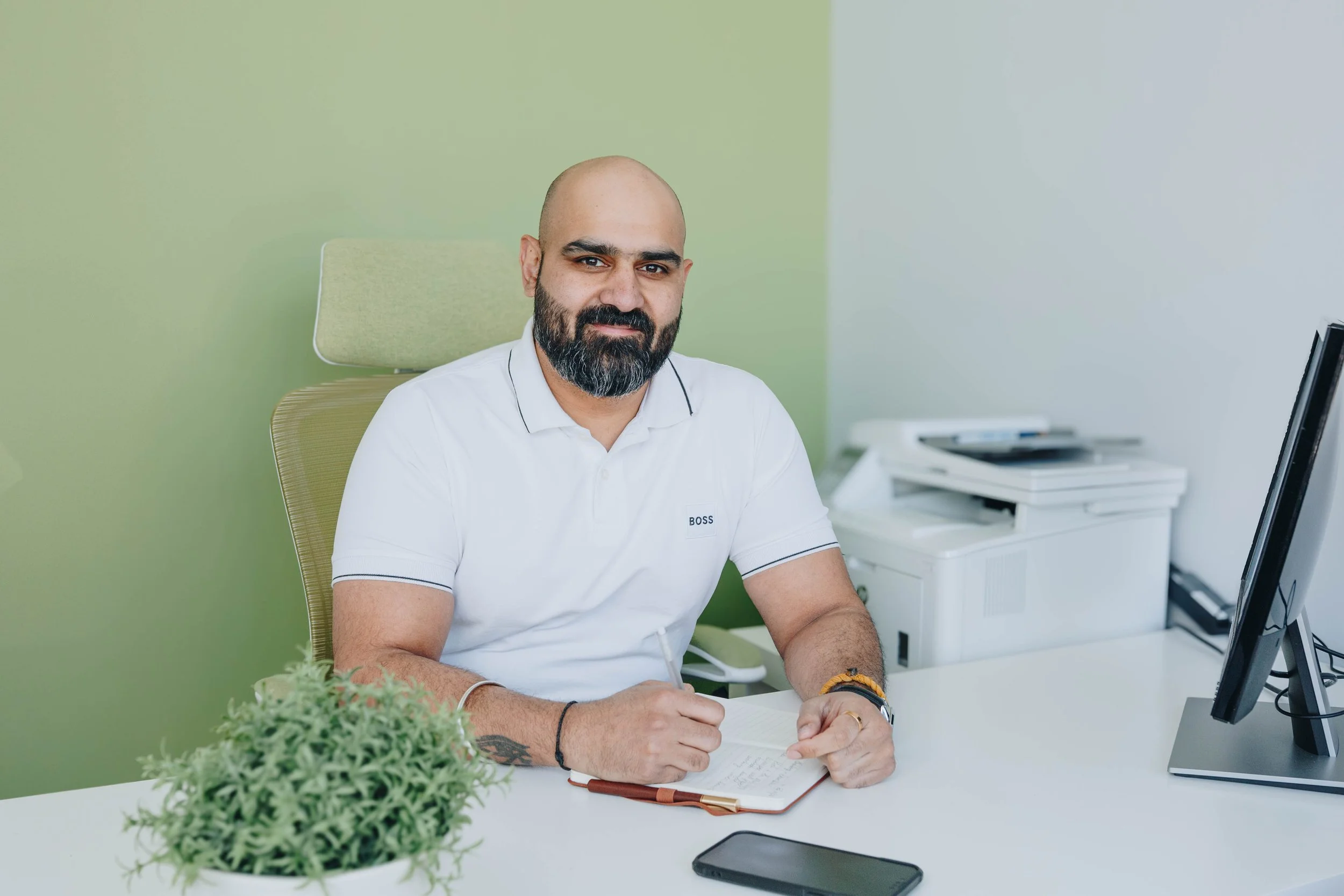 A man with a bald head and beard, wearing a white polo shirt, sitting at a desk with a notepad and pen, in an office with light green and white walls, and a computer monitor.